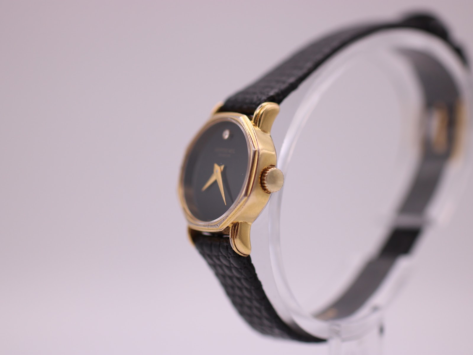 Angled side view of the Raymond Weil watch showing the gold-tone case and crown, on a clear display stand.