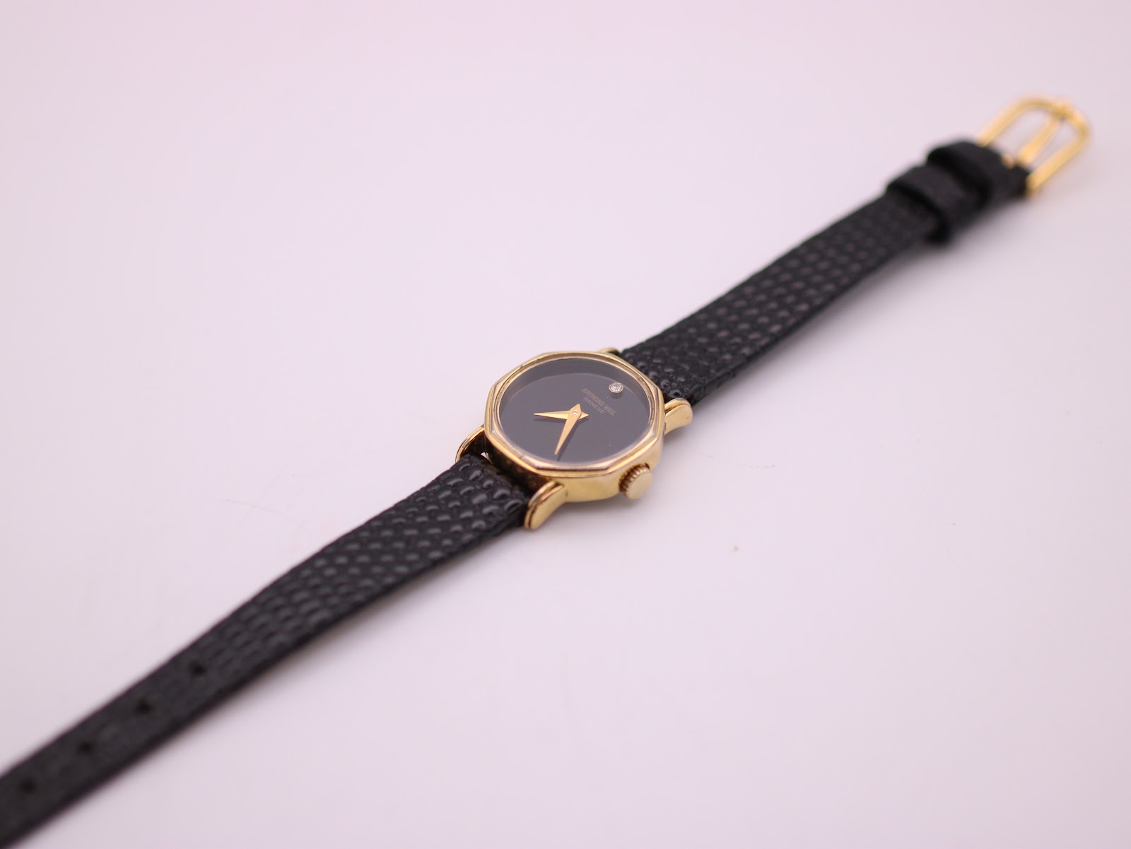 Overhead view of the full Raymond Weil watch, detailing the black textured leather strap and gold-tone buckle.