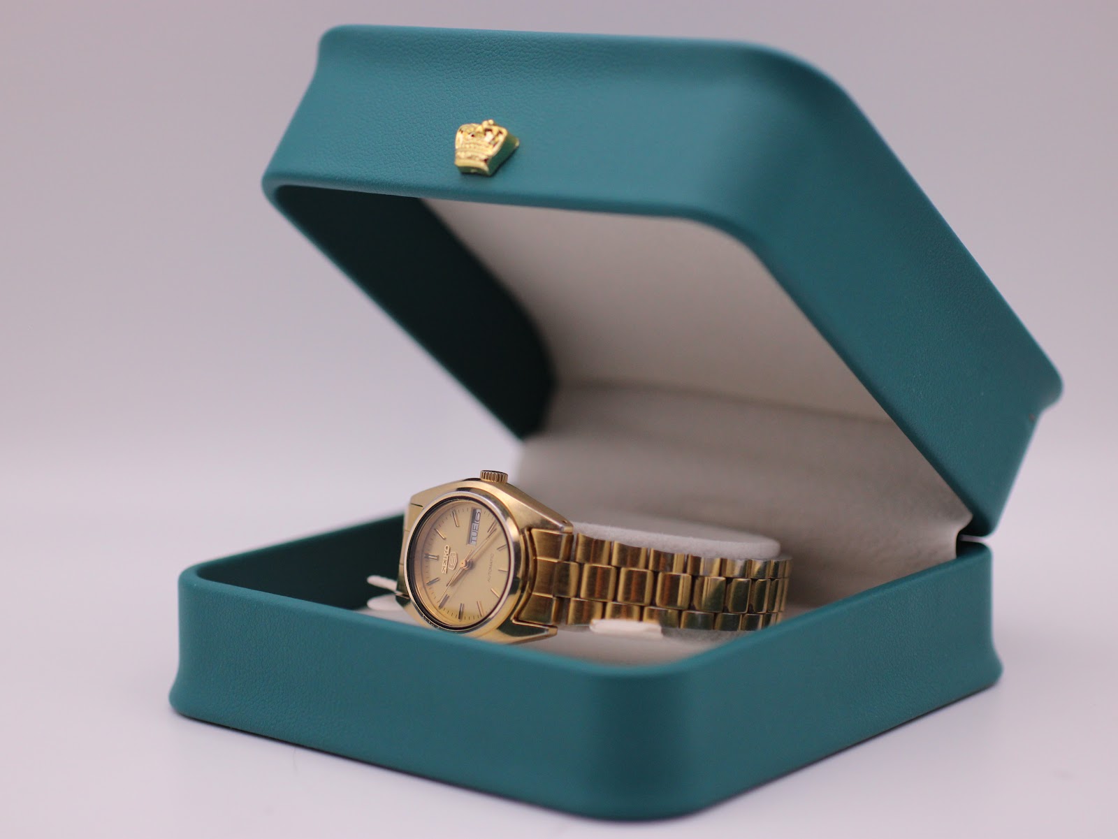 The gold-tone Seiko 5 watch presented in its original teal-coloured watch box with a gold crown emblem.