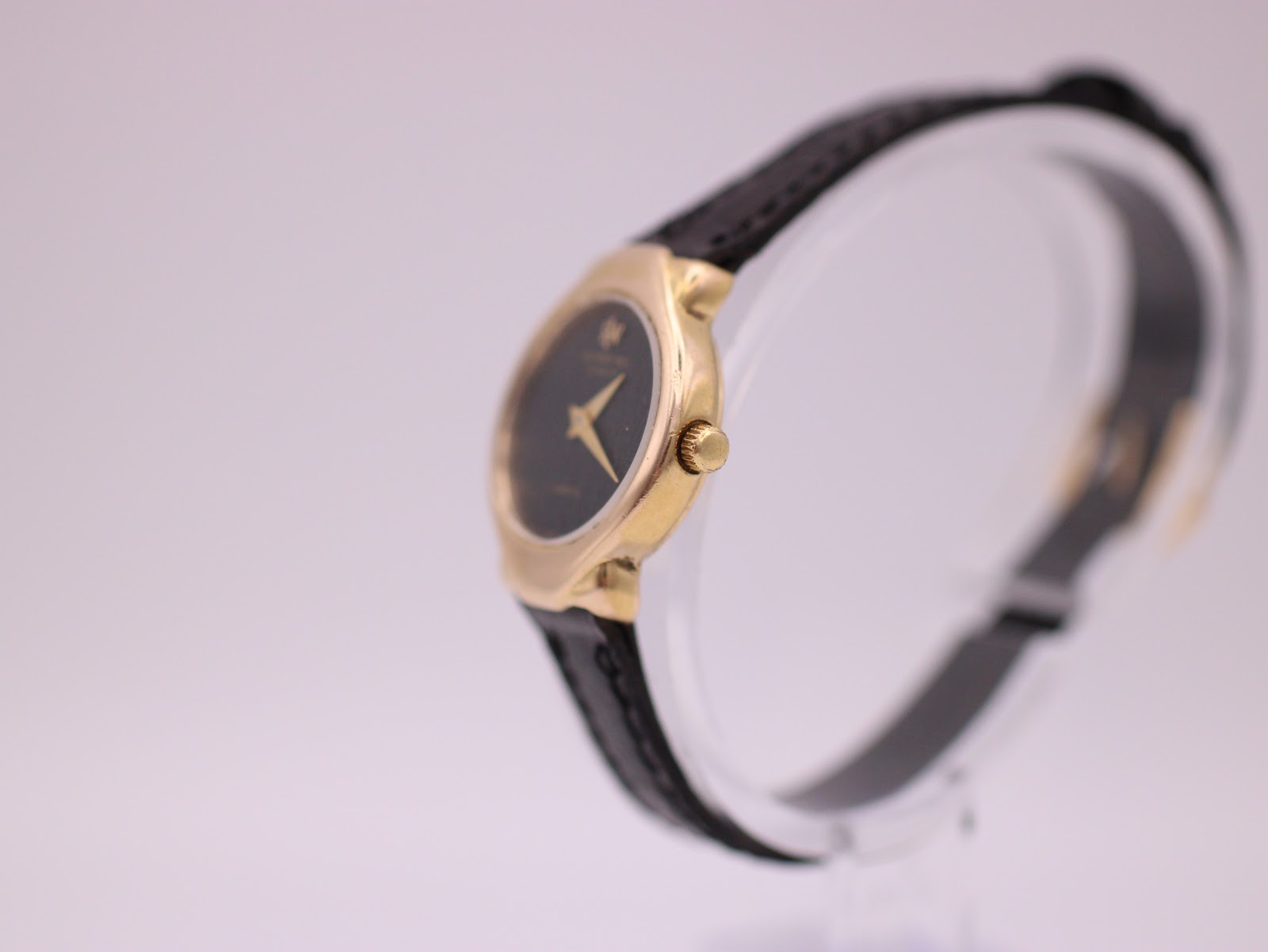 Side profile of the Raymond Weil watch showing the gold case, fluted crown, and black leather strap on a clear display stand.