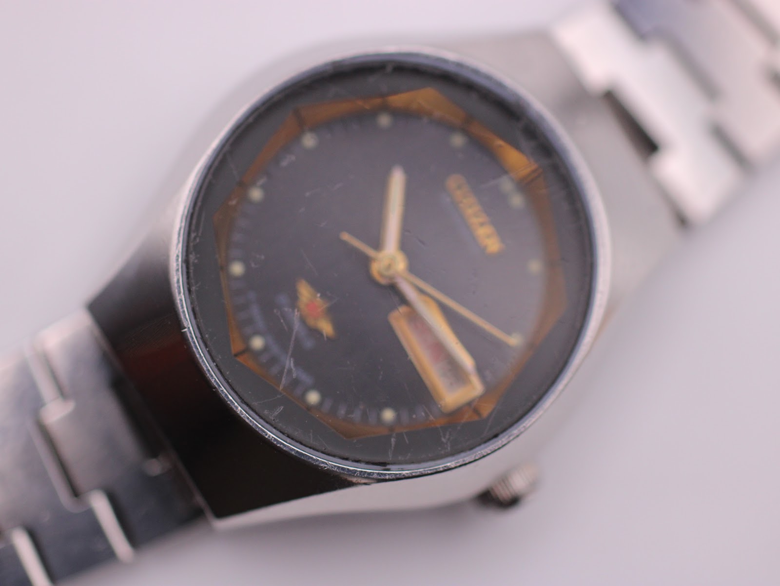 Close-up of the Citizen watch dial, showing scratches on the crystal, gold-tone hands, and day-date window.