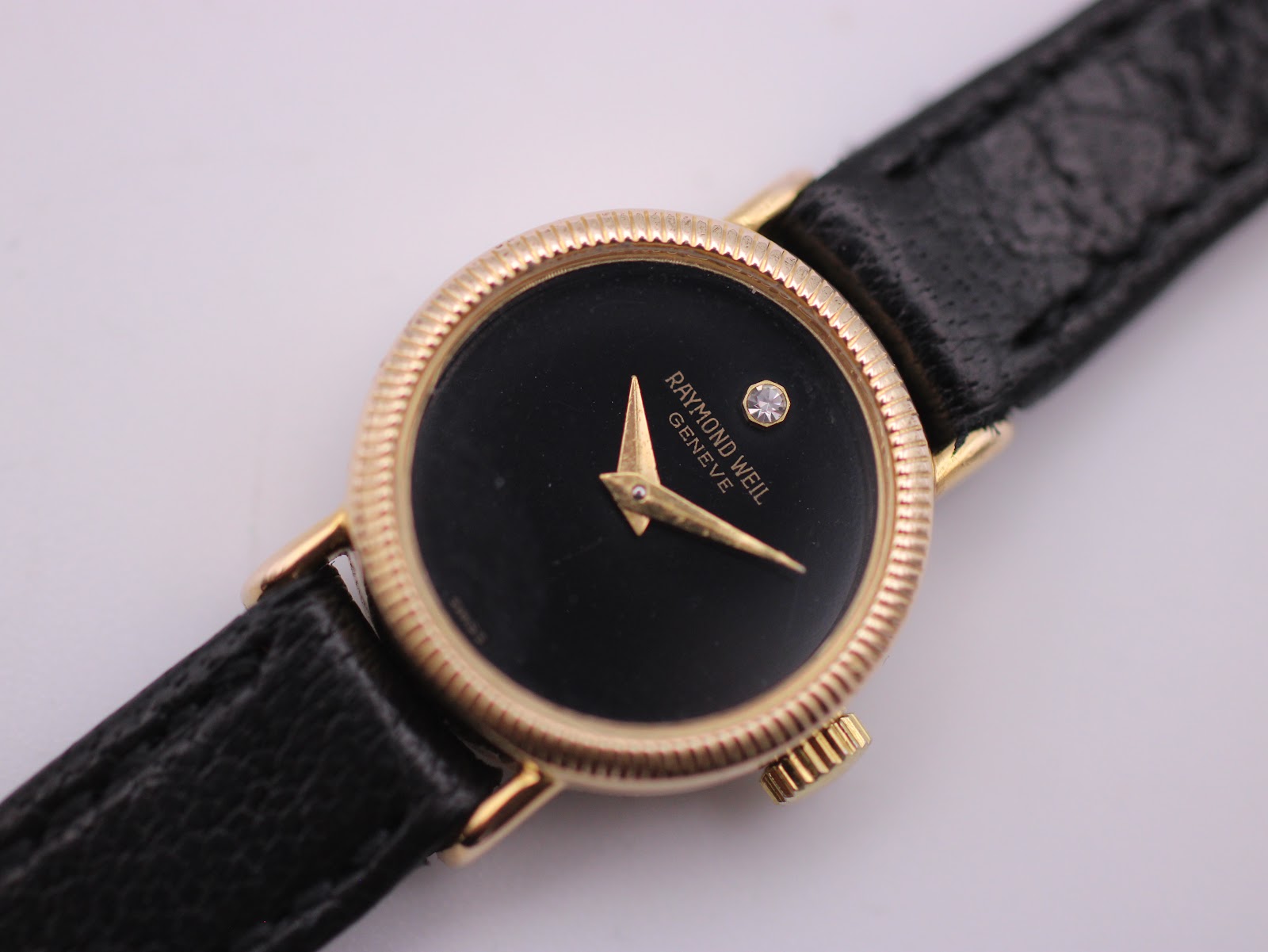 Close-up of the Raymond Weil black dial with gold tone hands and diamond marker at 12 o'clock.