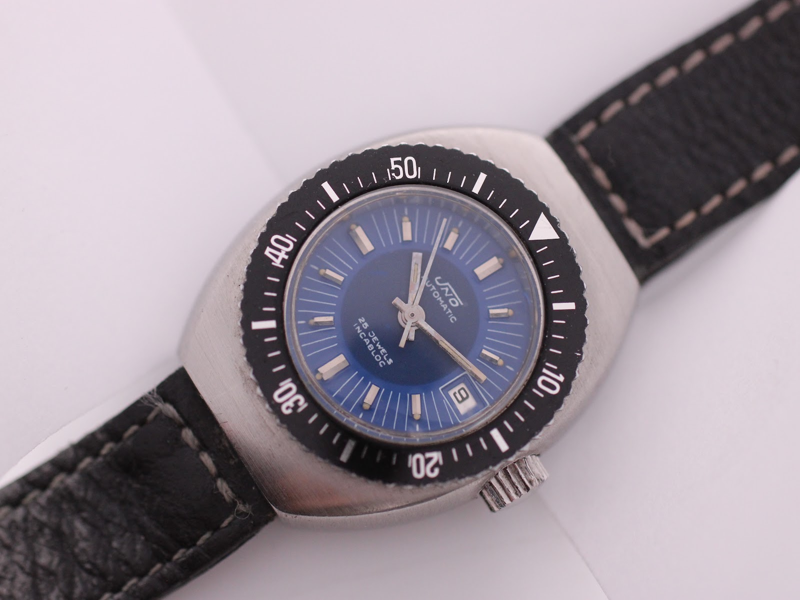 Angled close-up of the UNTO watch dial, highlighting the blue colour, applied indices, and date window.
