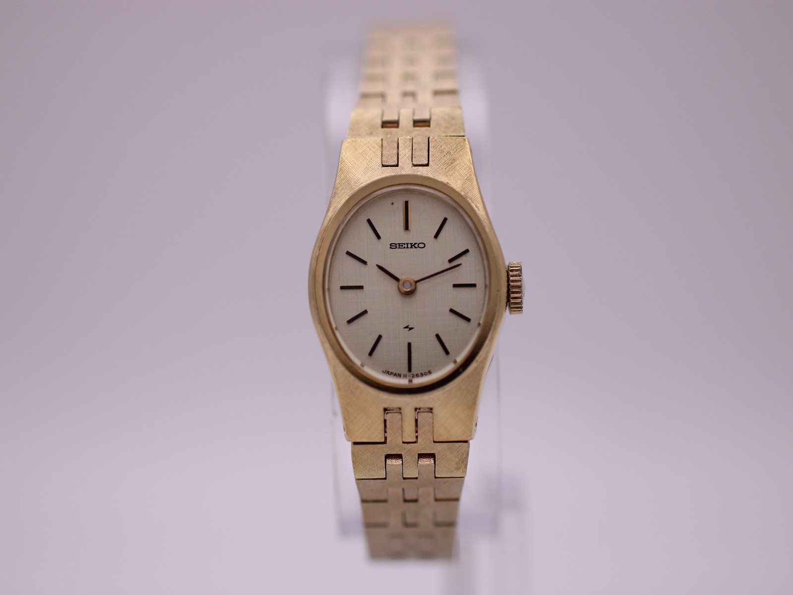 Front view of the gold-tone Seiko ladies watch with a silver textured dial and integrated bracelet, on a clear stand.