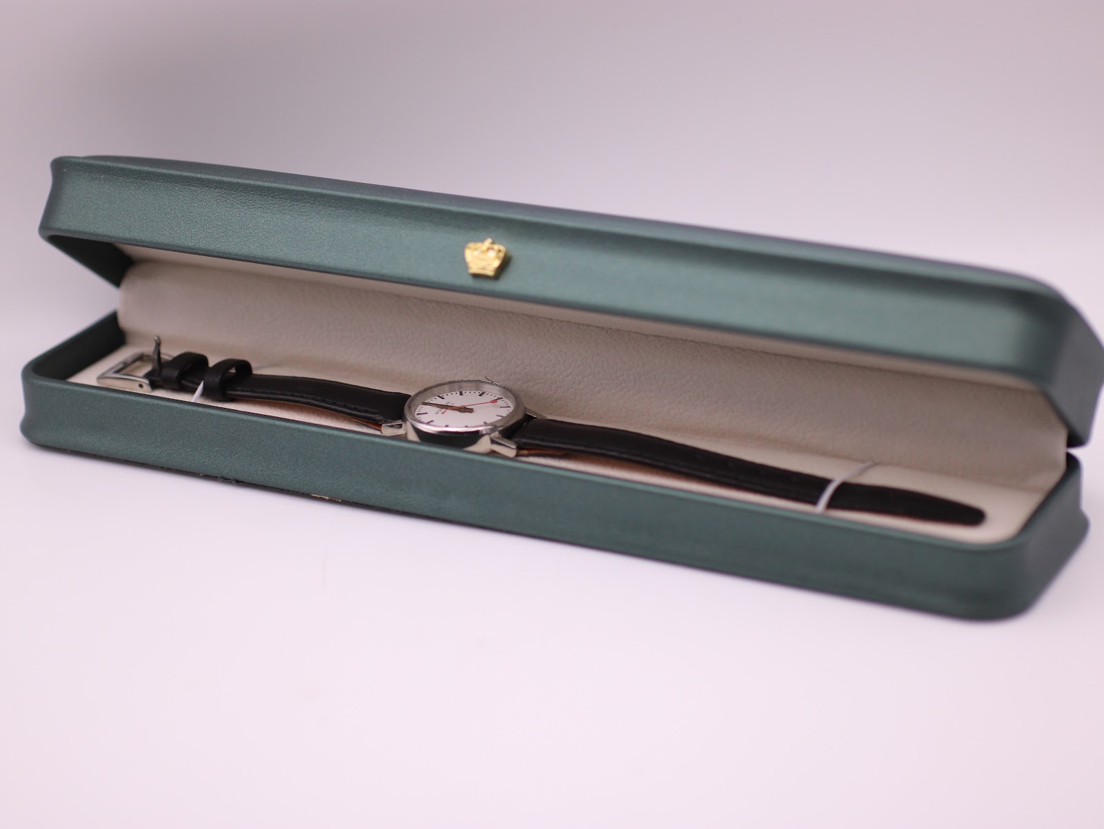 The Mondaine watch resting inside a generic green leatherette presentation box with a gold crown emblem.