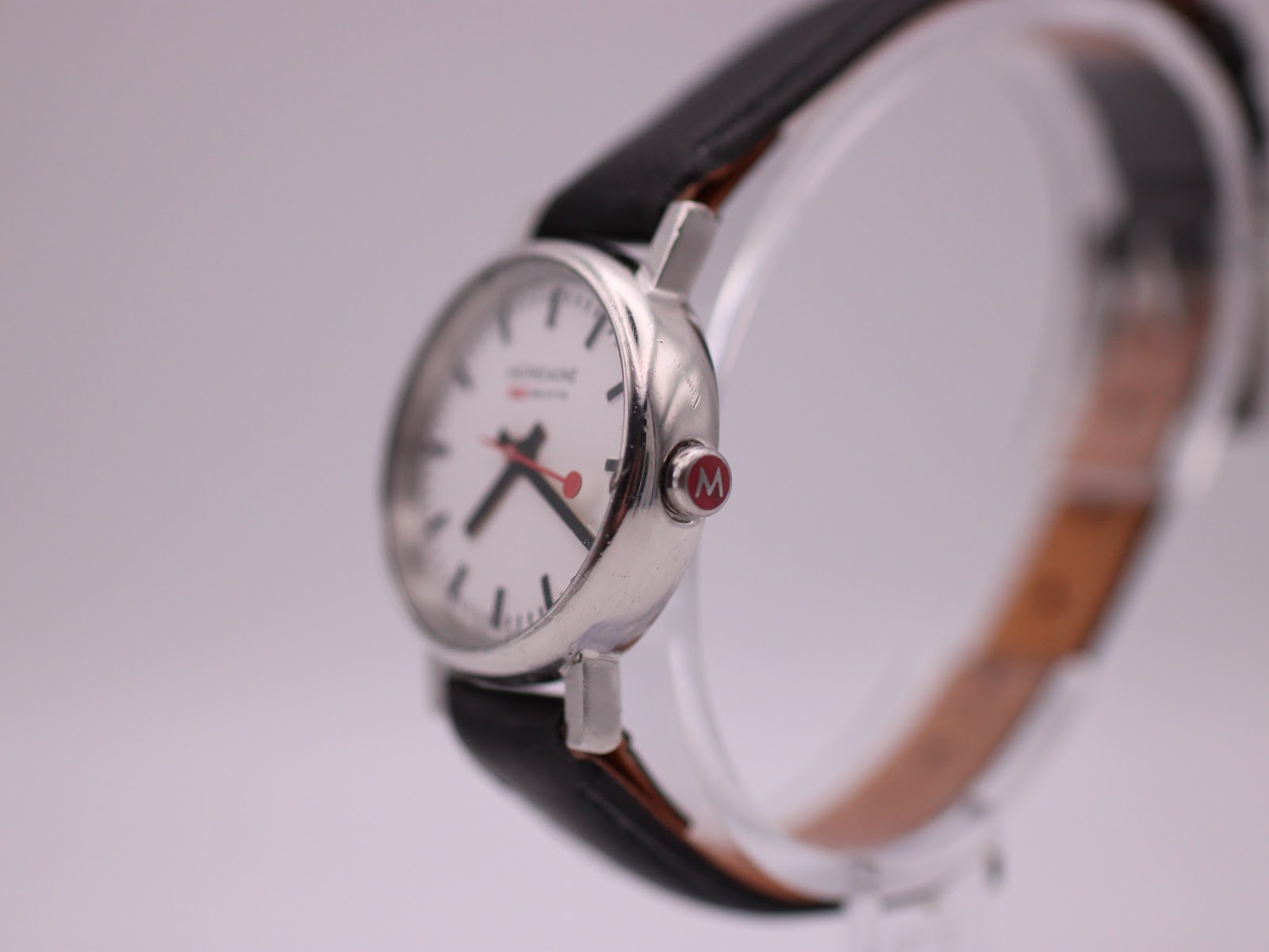 Angled side view of the Mondaine watch showing the polished silver case, red 'M' crown, and black leather strap.