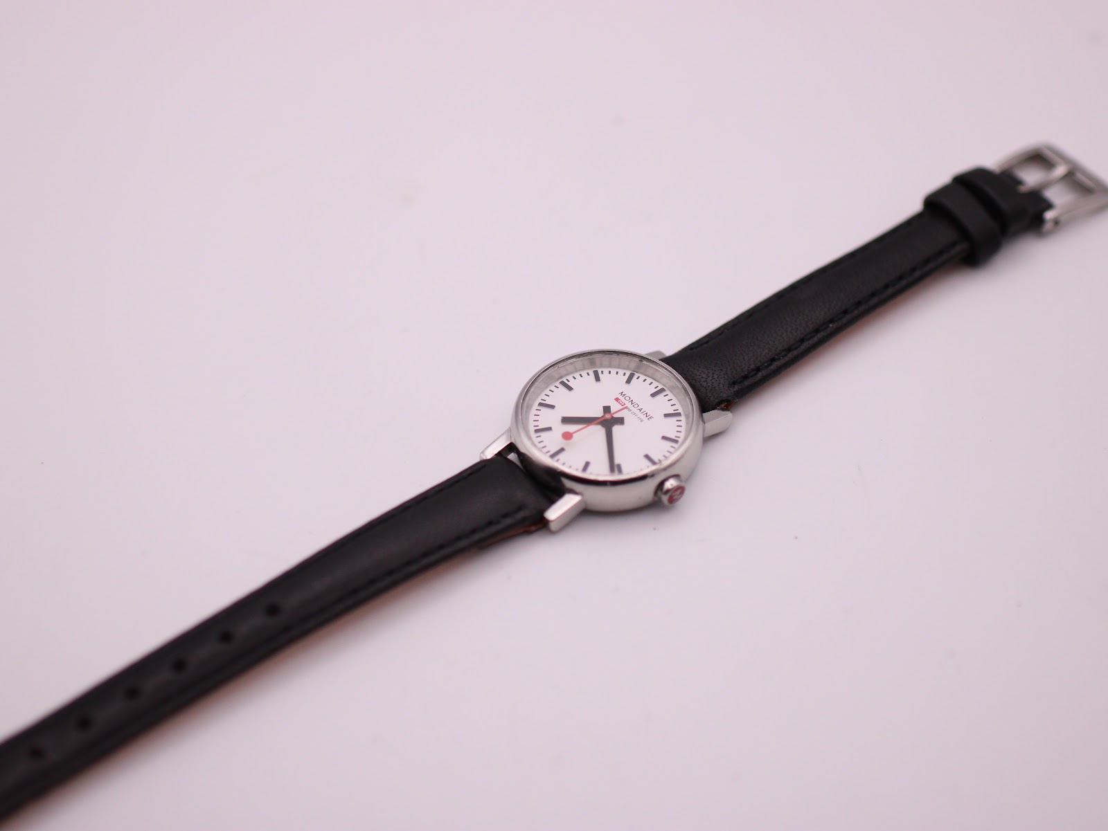 Overhead angled view of the entire Mondaine watch, laid out on a white background, showing the full length of the black leather strap.