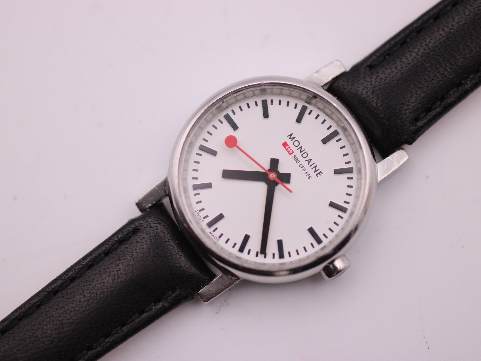 Close-up front view of the Mondaine white dial with clear black baton hour markers and the iconic red seconds hand, partially showing the black leather strap.