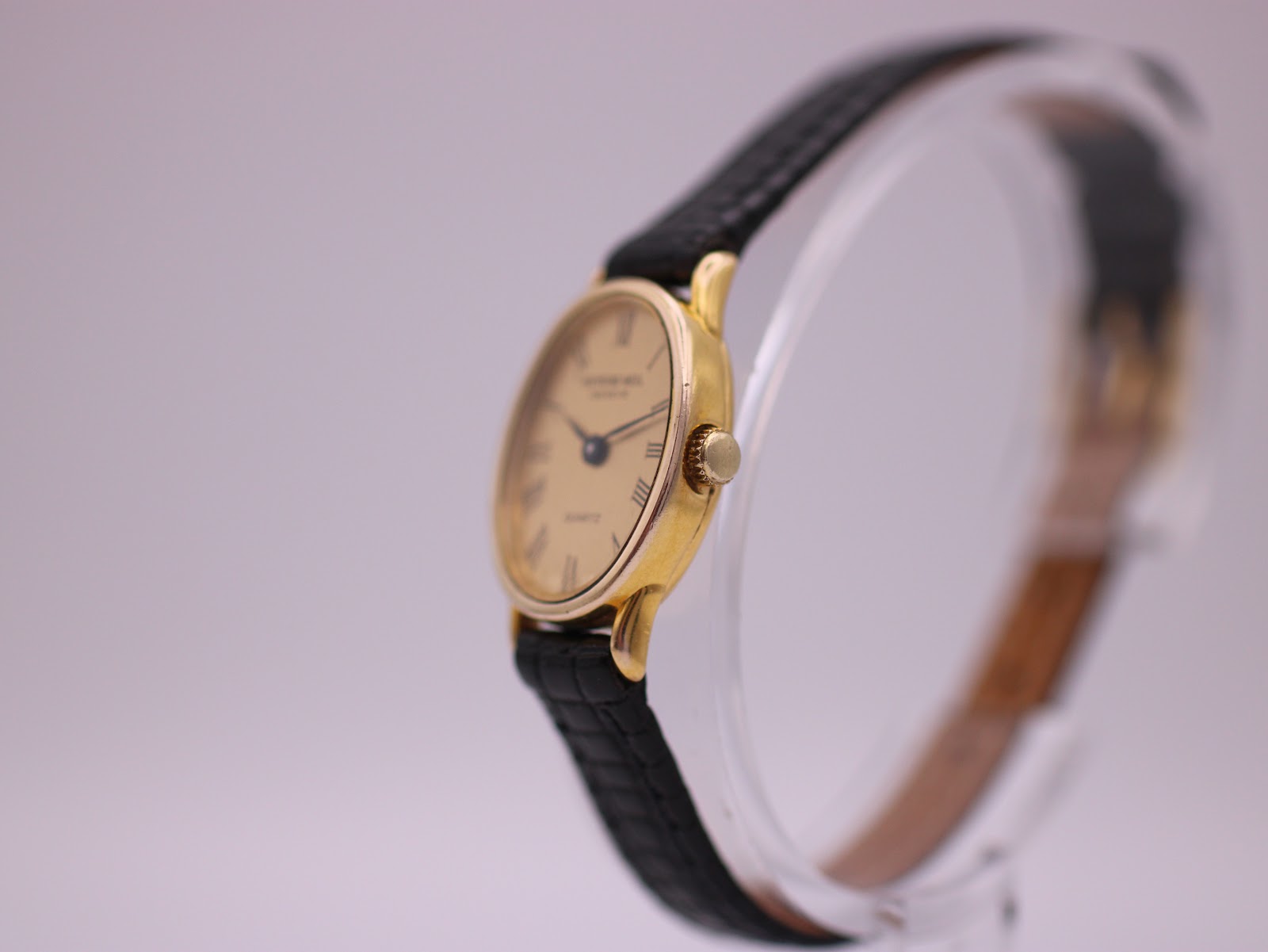 Angled side view of the Raymond Weil watch showing the gold electroplated case and crown, with the black leather strap curving back.