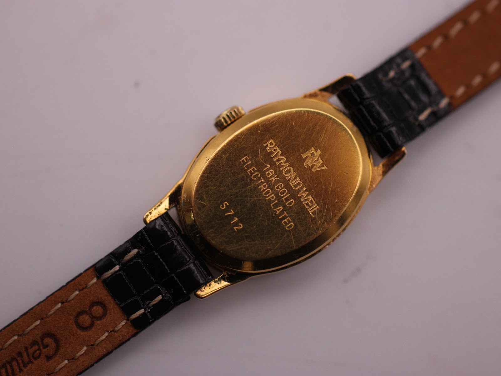 Close-up of the watch case back with 'RAYMOND WEIL 18K GOLD ELECTROPLATED 5712' engraving, and the strap showing 'Genuine Leather 8' markings.