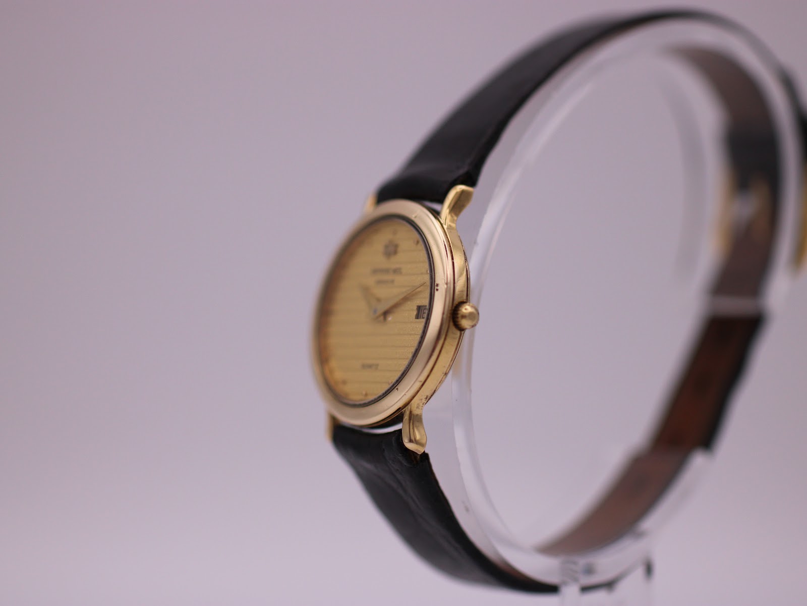 Side angled view of the Raymond Weil watch showing the gold electroplated case and crown, displayed on a clear stand.