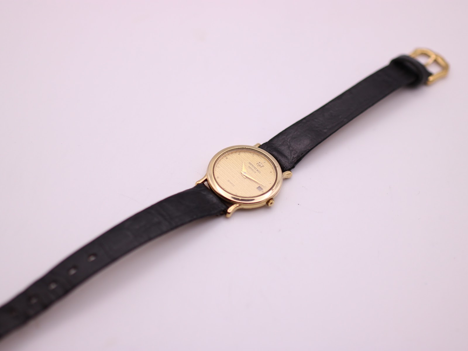 Overhead full view of the Raymond Weil watch with black leather strap and gold tone buckle on a white background.