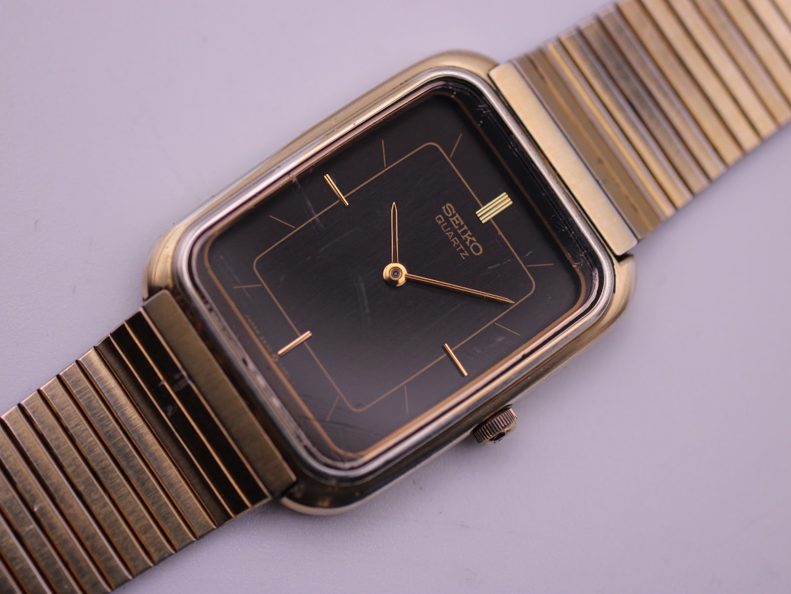 Close-up of the black dial of the Seiko watch, showing gold baton indices, hands, and visible crystal crack.