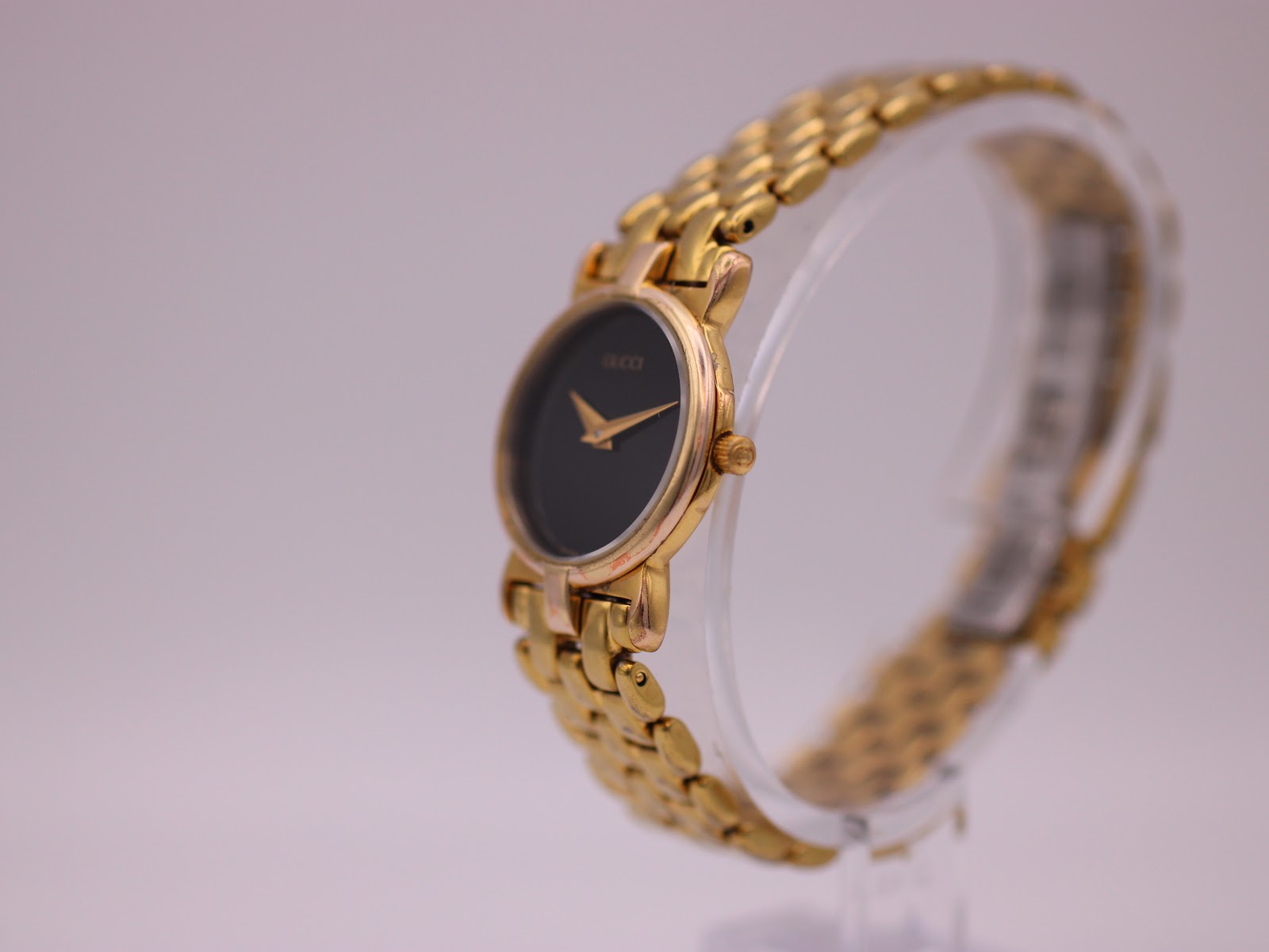 Angled left view of the Gucci 3400 L watch on a clear display stand, highlighting the gold-tone case and bracelet.