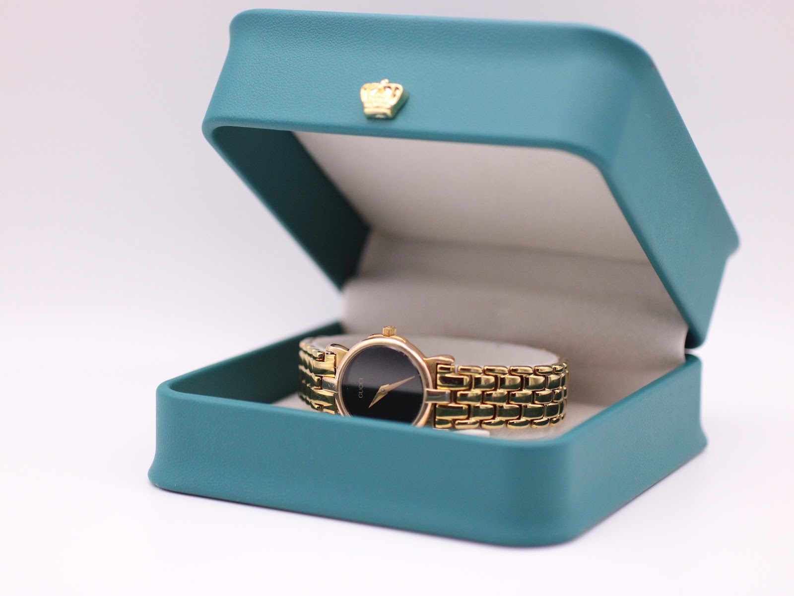 The Gucci watch displayed open inside a generic green presentation box with a gold crown emblem, a studio prop.