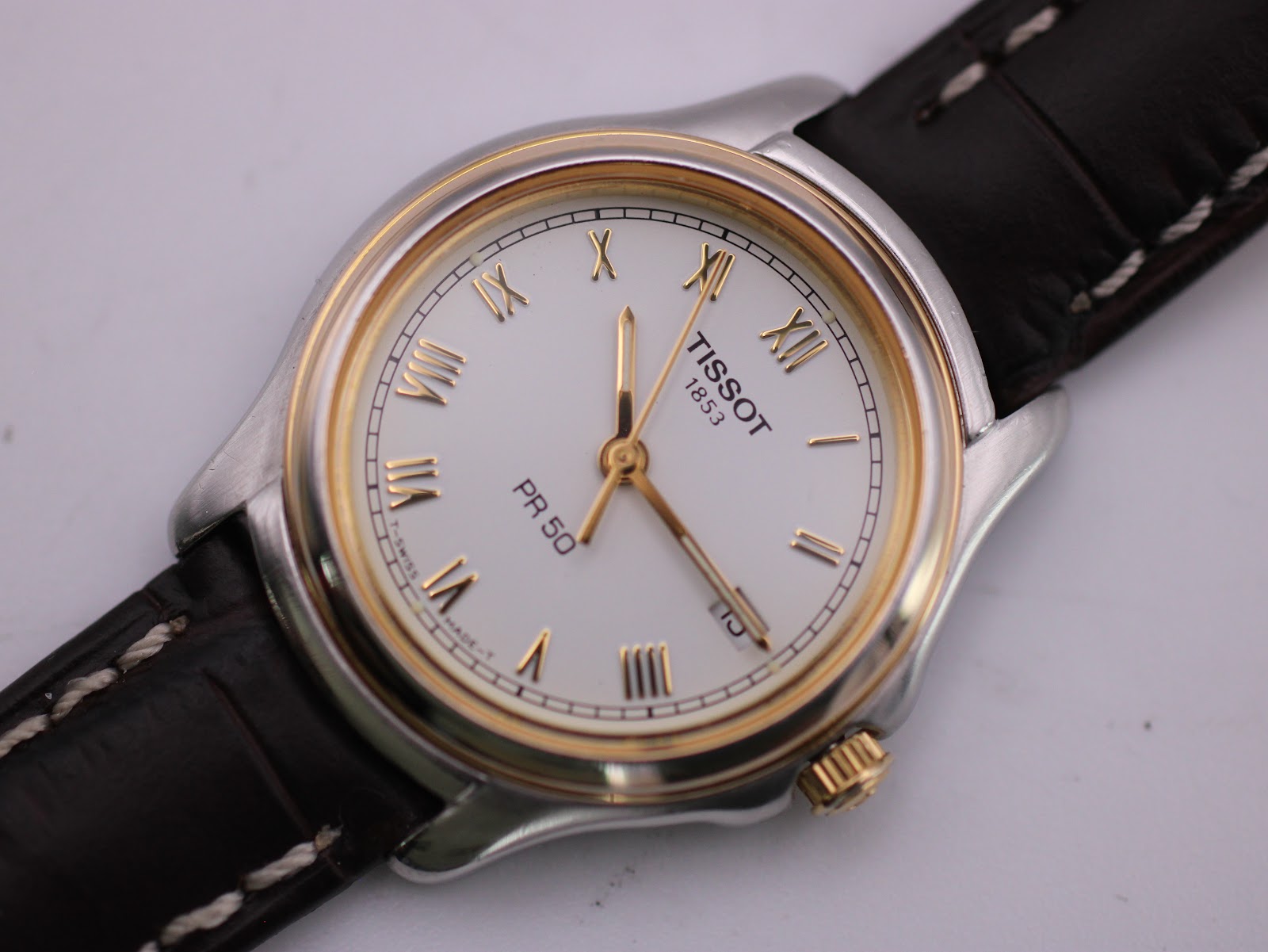 Close-up of the Tissot PR 50's white dial, featuring gold-tone Roman numeral hour markers and hands, with the Tissot logo and date window visible.