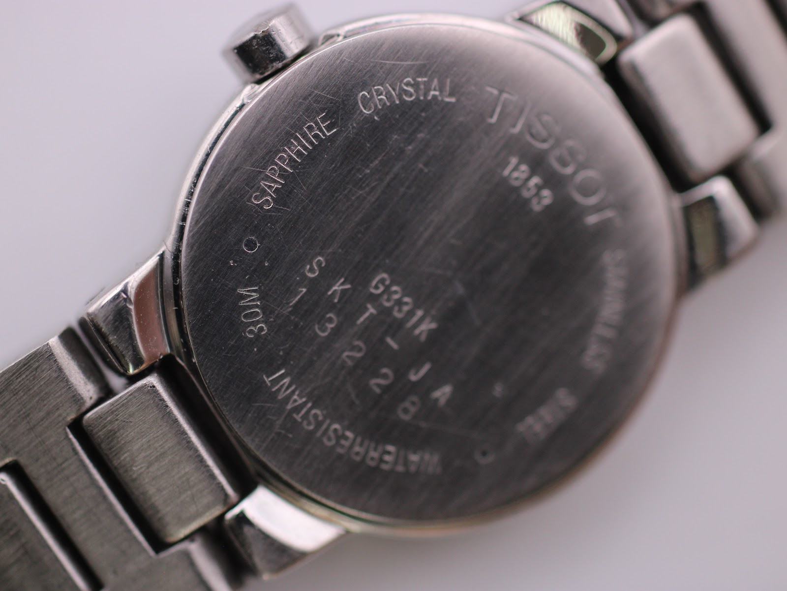 Close-up of the Tissot watch case back with engravings for 'Sapphire Crystal', '30M Water Resistant', 'Stainless Steel', 'G331K', and serial number details.