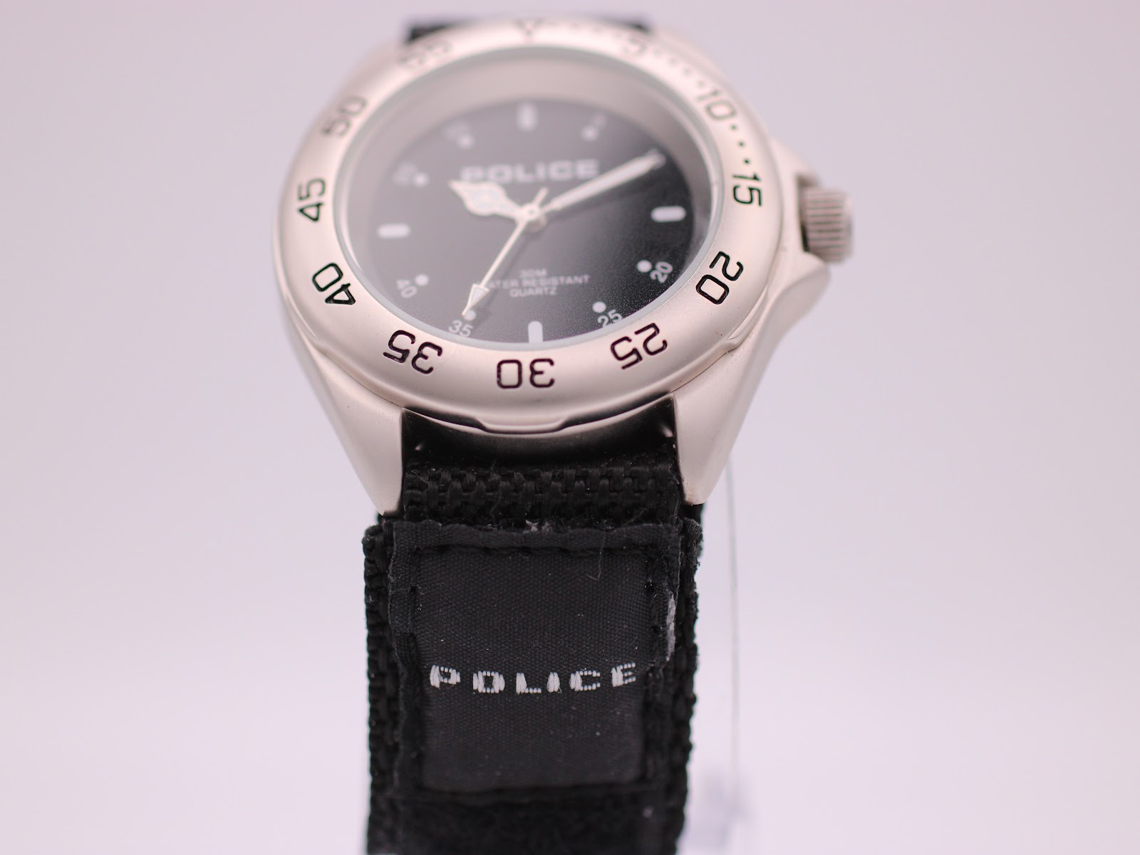 Frontal view of the Police watch, showing the black dial, silver case, and a close-up of the 'POLICE' branded tag on the fabric strap.
