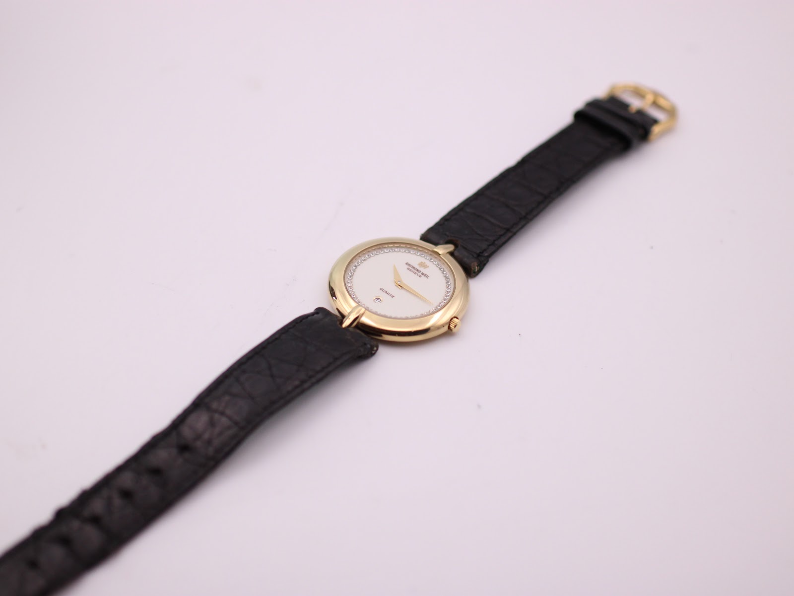 Top-down view of Raymond Weil 5350 watch with gold case and full black leather strap on a white background.