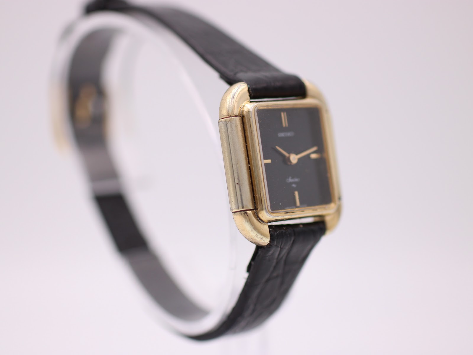 Angled three-quarter view of the Seiko Socie watch, highlighting the gold-plated case and black dial on a clear stand.