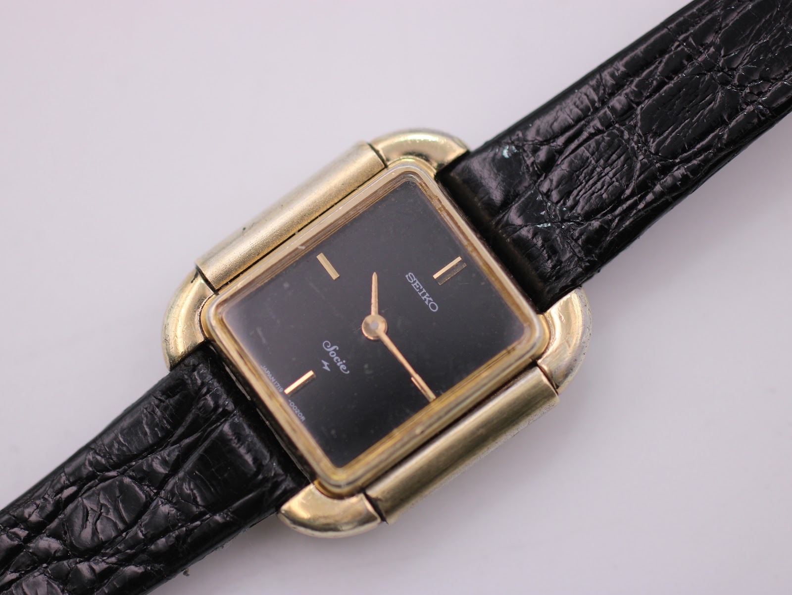 Close-up detailed view of the black dial of the Seiko Socie, showing the "Seiko" and "Socie" text, gold baton indices, and hands.