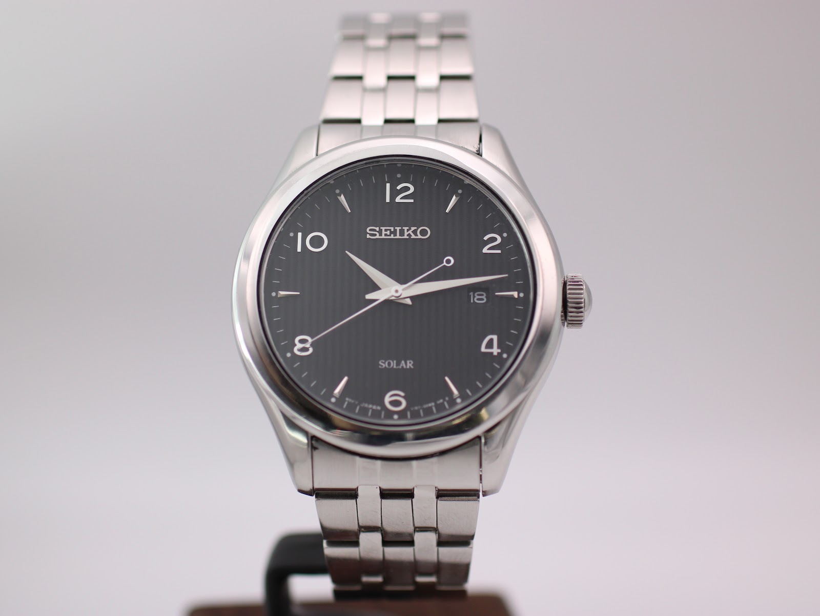 Front view of Seiko V157-0CNO solar watch with black vertically textured dial and stainless steel bracelet.