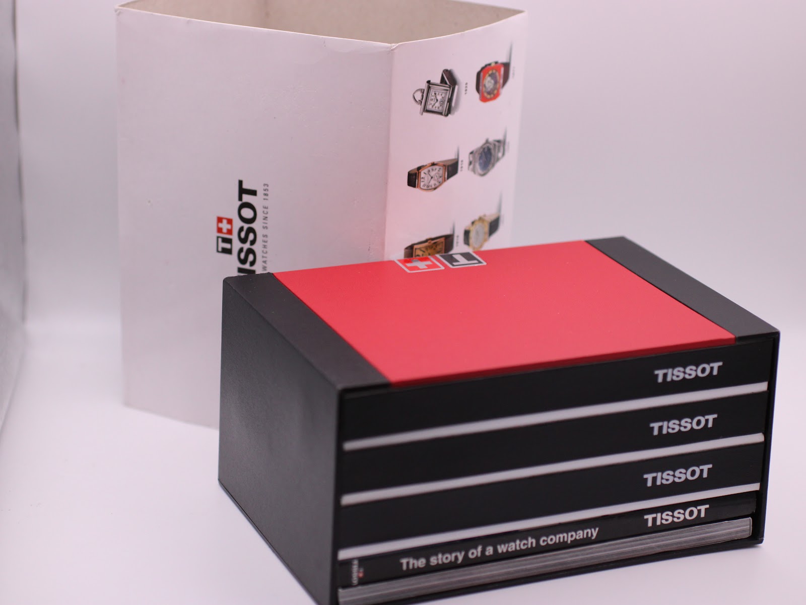 The red and black original Tissot presentation box with a white outer sleeve and a 'The story of a watch company' booklet.