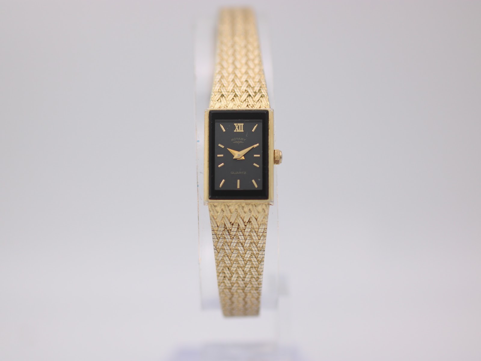 Front view of the gold-tone Rotary ladies watch with a black rectangular dial on a clear stand.