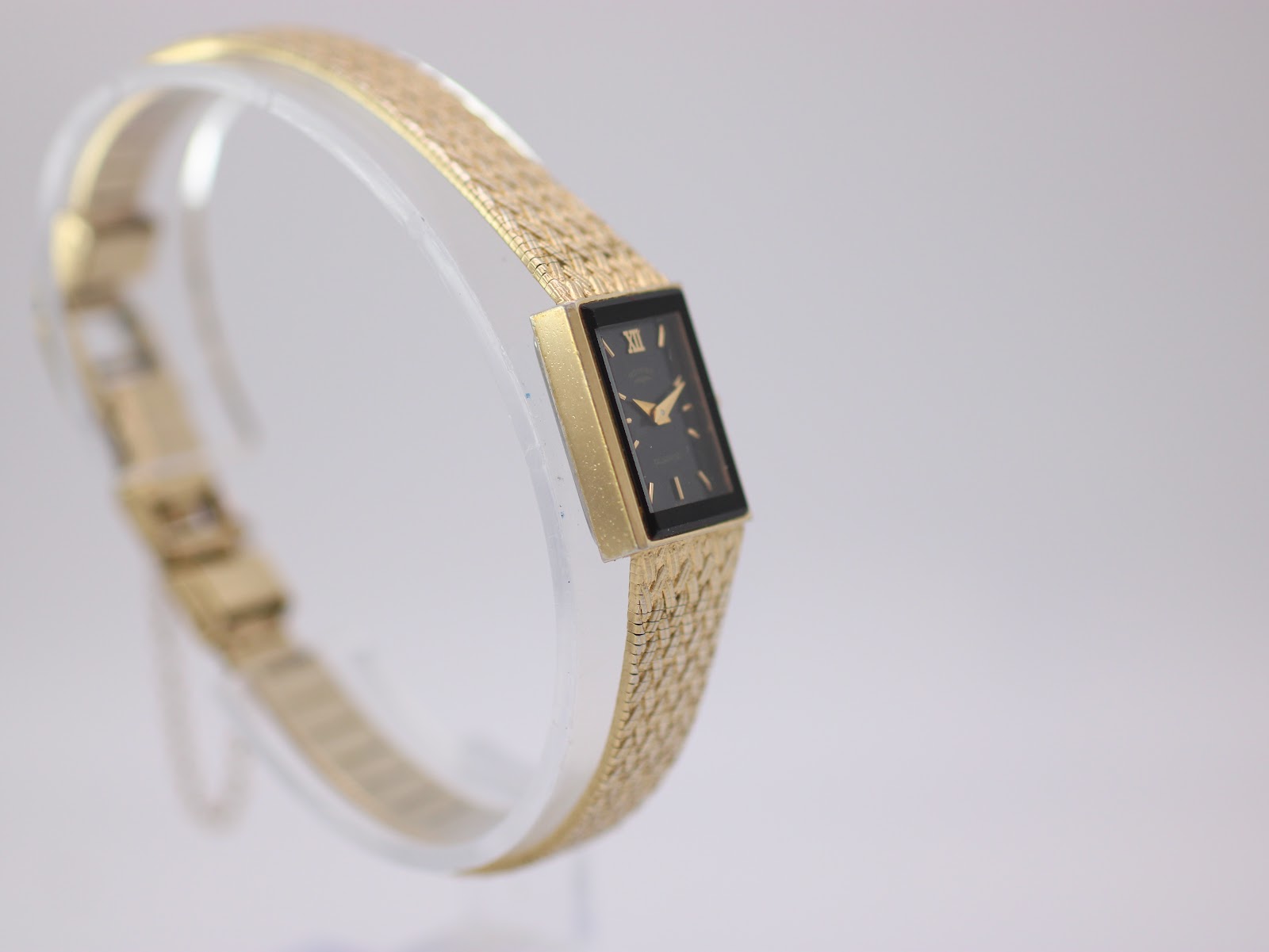 Angled side view of the Rotary watch, showing the gold-tone rectangular case and mesh bracelet on a clear stand.
