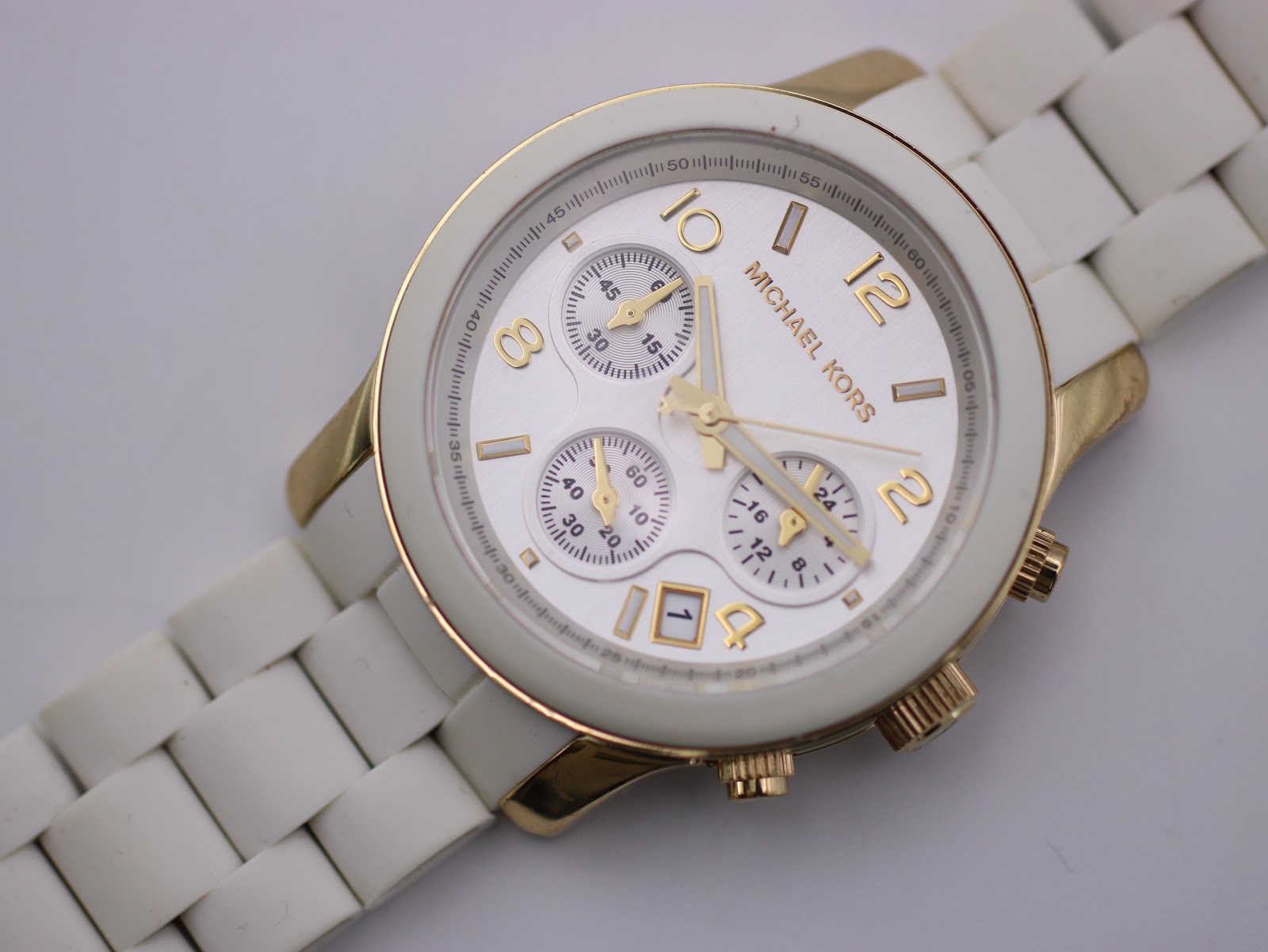 Close-up, angled view of the Michael Kors white chronograph dial with gold-tone details and the white ceramic bracelet extending away.