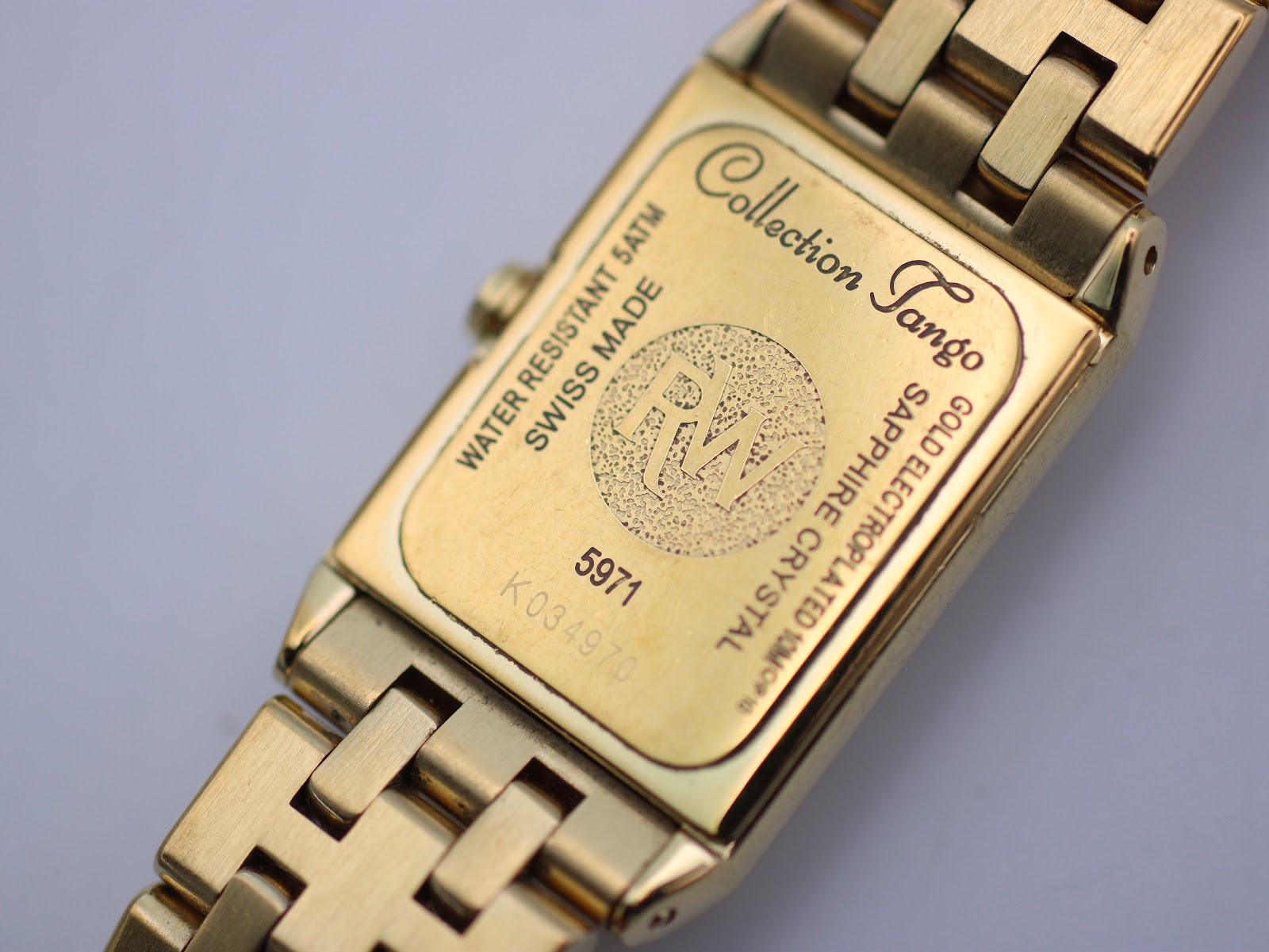 Close-up of the watch case back, detailing 'Collection Tango', model number 5971, serial K034970, 'Water Resistant 5ATM', and 'Sapphire Crystal'.