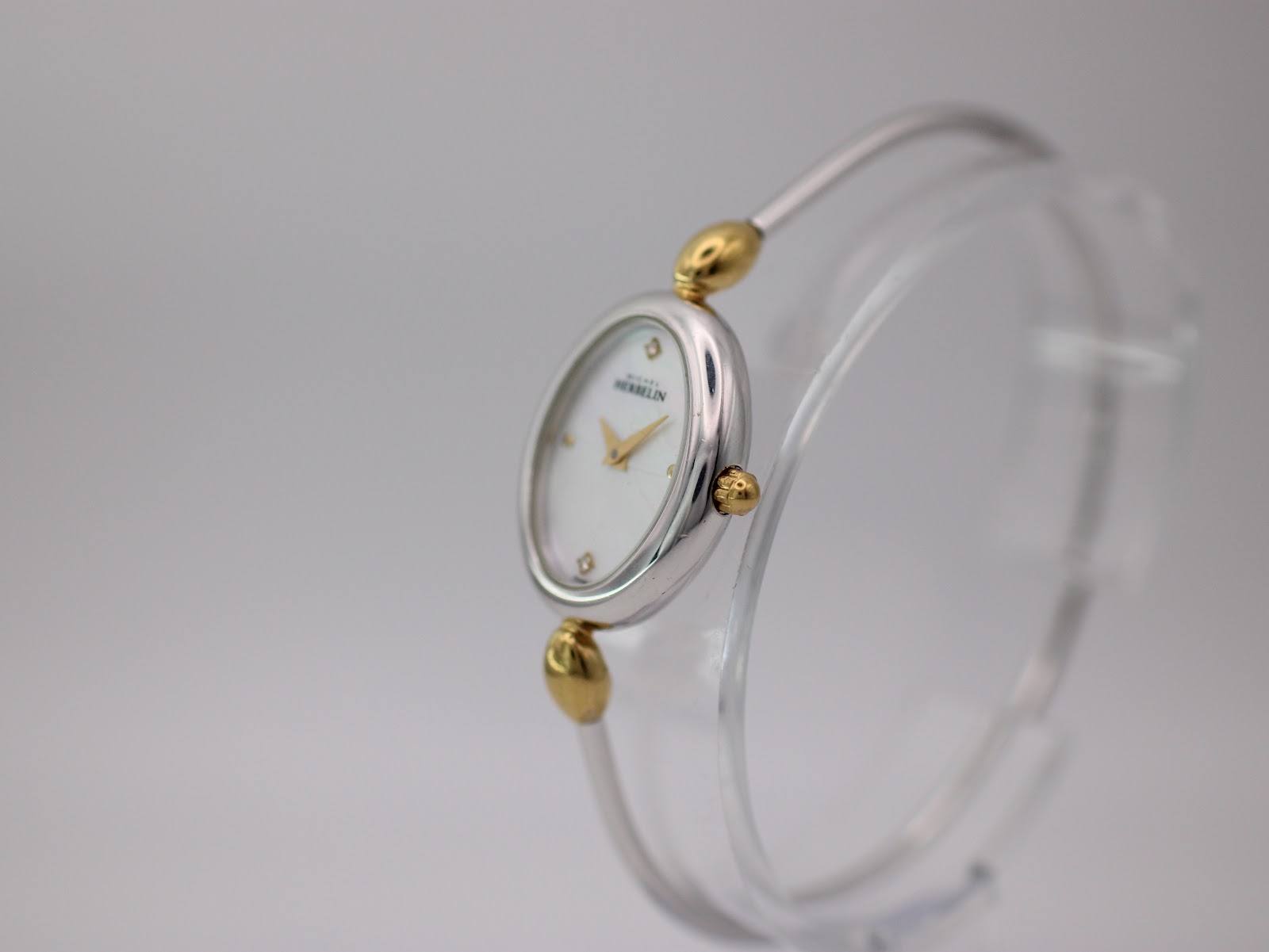 Angled side view of the Michel Herbelin watch on a clear stand, showing the oval case and integrated two-tone bangle.