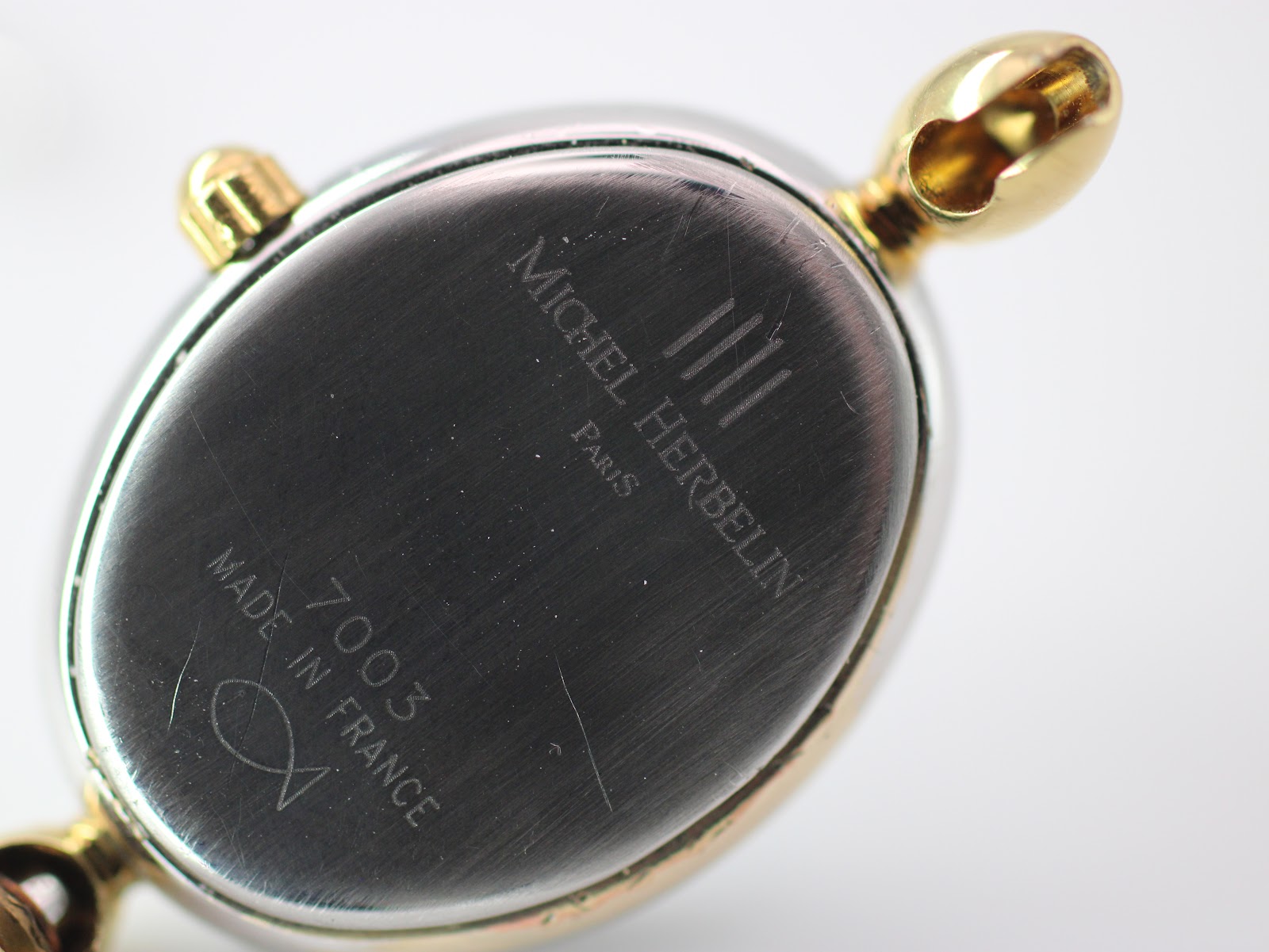 Detailed view of the watch case back, showing 'Michel Herbelin Paris' and '7003 Made In France' engravings with surface scratches.