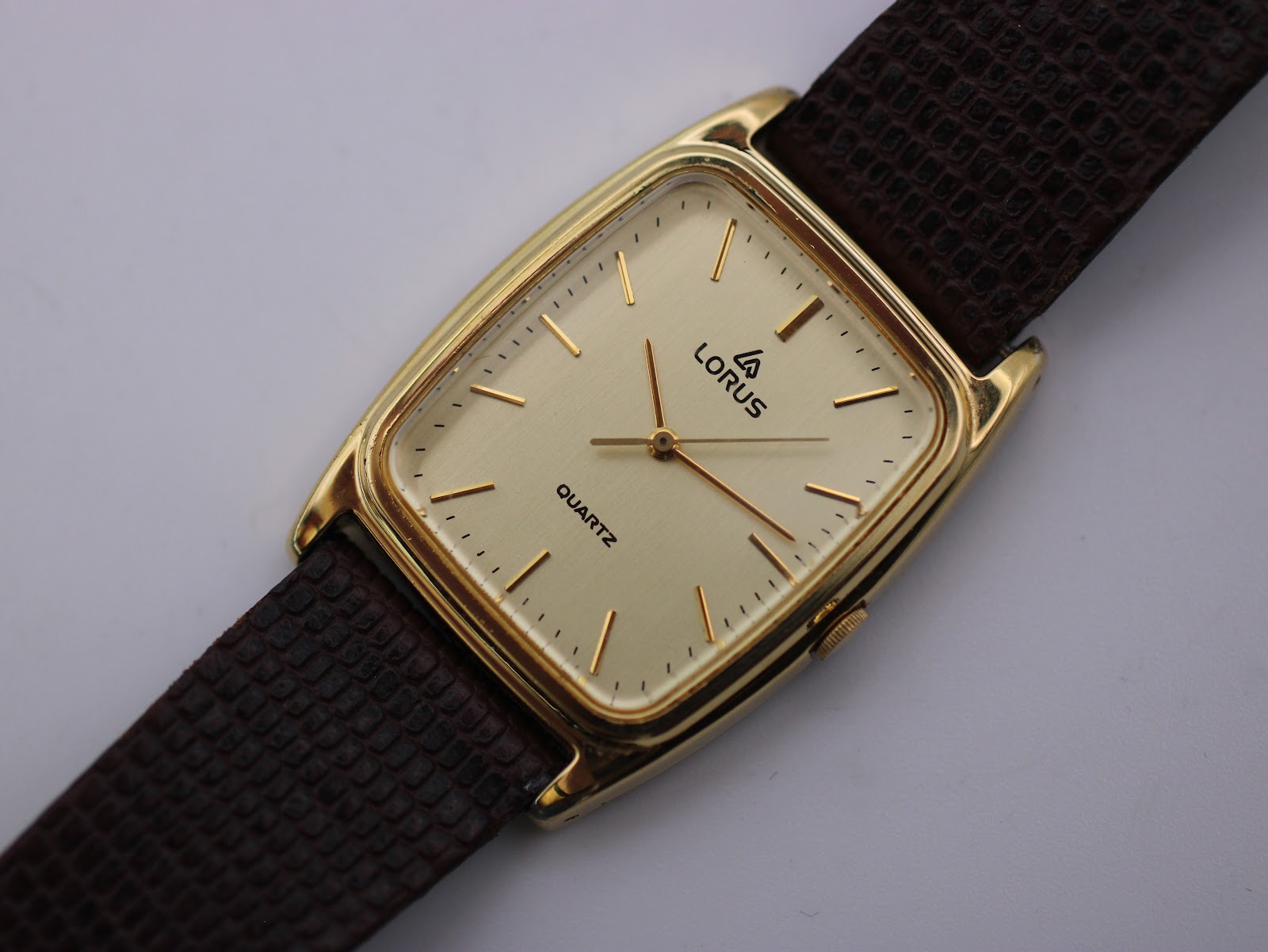 Close-up, slightly elevated view of the Lorus watch dial, showing the Lorus logo, 'QUARTZ' text, gold-tone hands, and the textured brown strap.