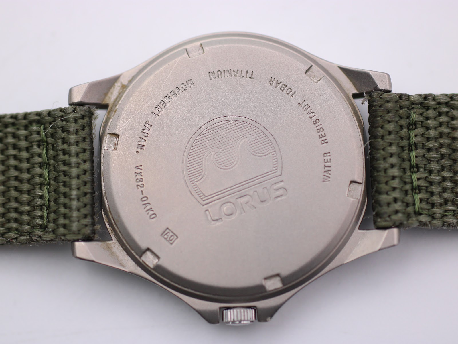 Close-up of Lorus watch case back, showing 'TITANIUM', 'WATER RESISTANT 10BAR', 'MOVEMENT JAPAN', and model number VX82-0AY0.
