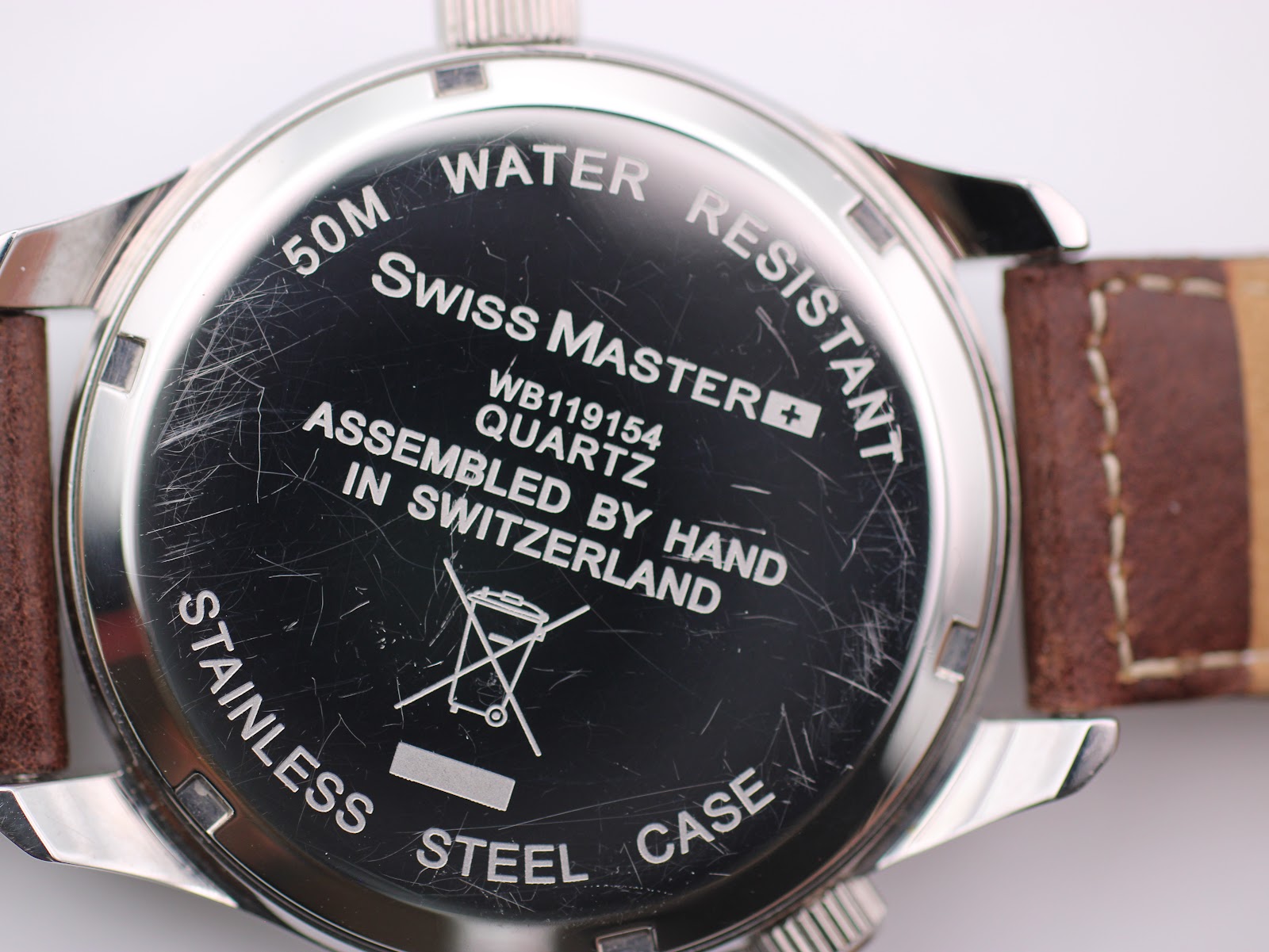 Macro close-up of the watch case back, displaying engravings '50M WATER RESISTANT', 'Swiss Master', 'WB119154', 'QUARTZ', 'ASSEMBLED BY HAND IN SWITZERLAND', and 'STAINLESS STEEL CASE', along with visible scratches.