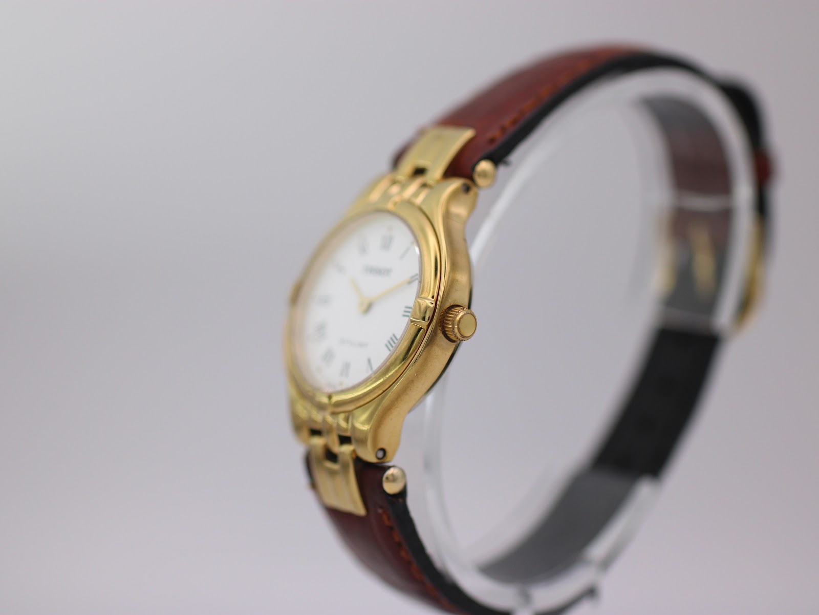 Angled side view of the Tissot Stylist on a display stand, showing the curved gold case and brown strap.