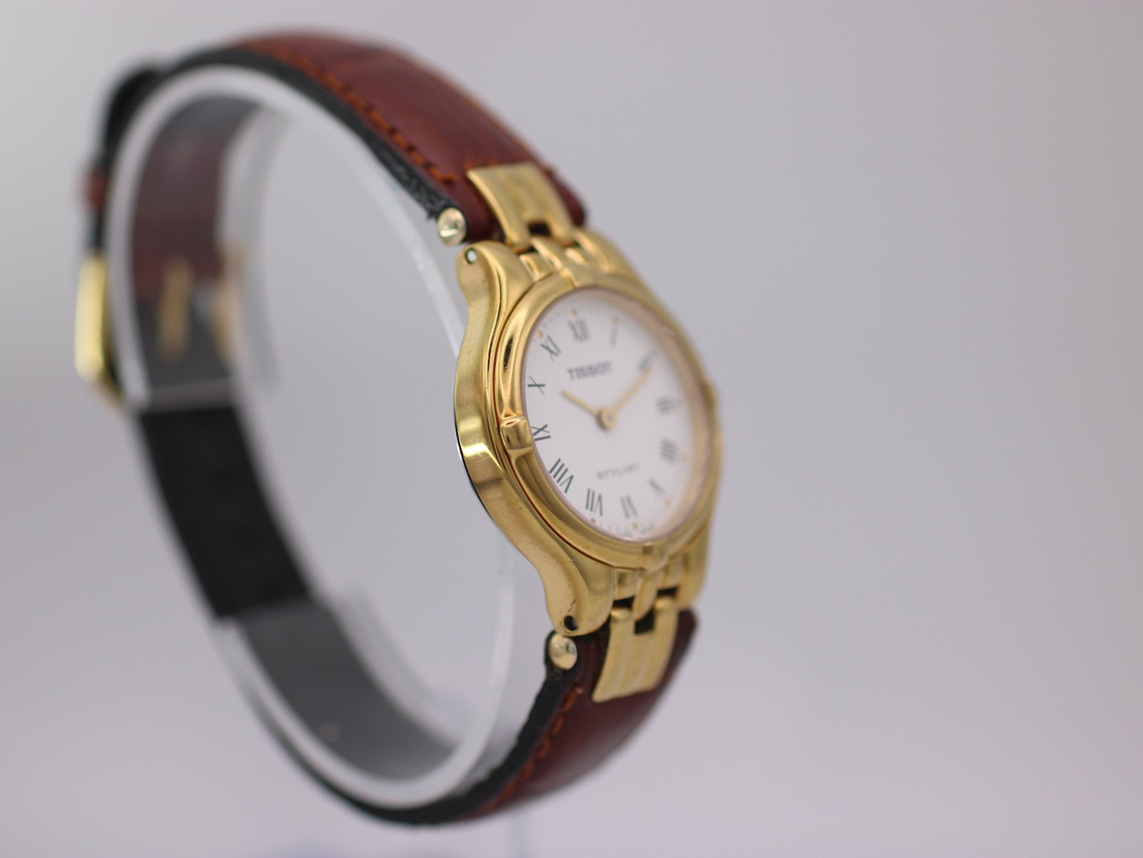 Opposite angled side view of the Tissot Stylist on a display stand, highlighting the gold-tone case profile.