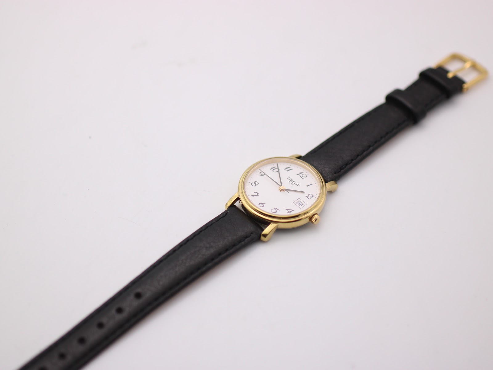 Top-down view of the Tissot watch with the black leather strap extended, showing the gold tone buckle.
