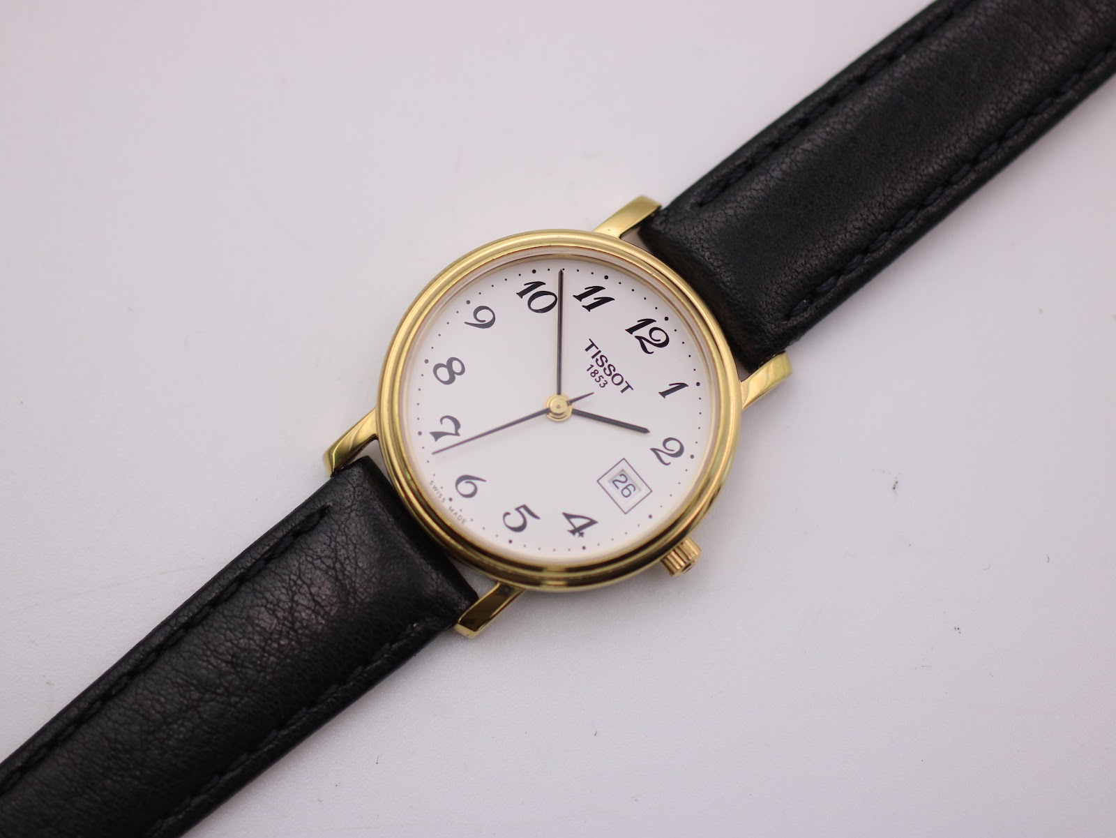 Close-up, slightly angled front view of the Tissot white dial with date window and gold hands, showing the full strap.