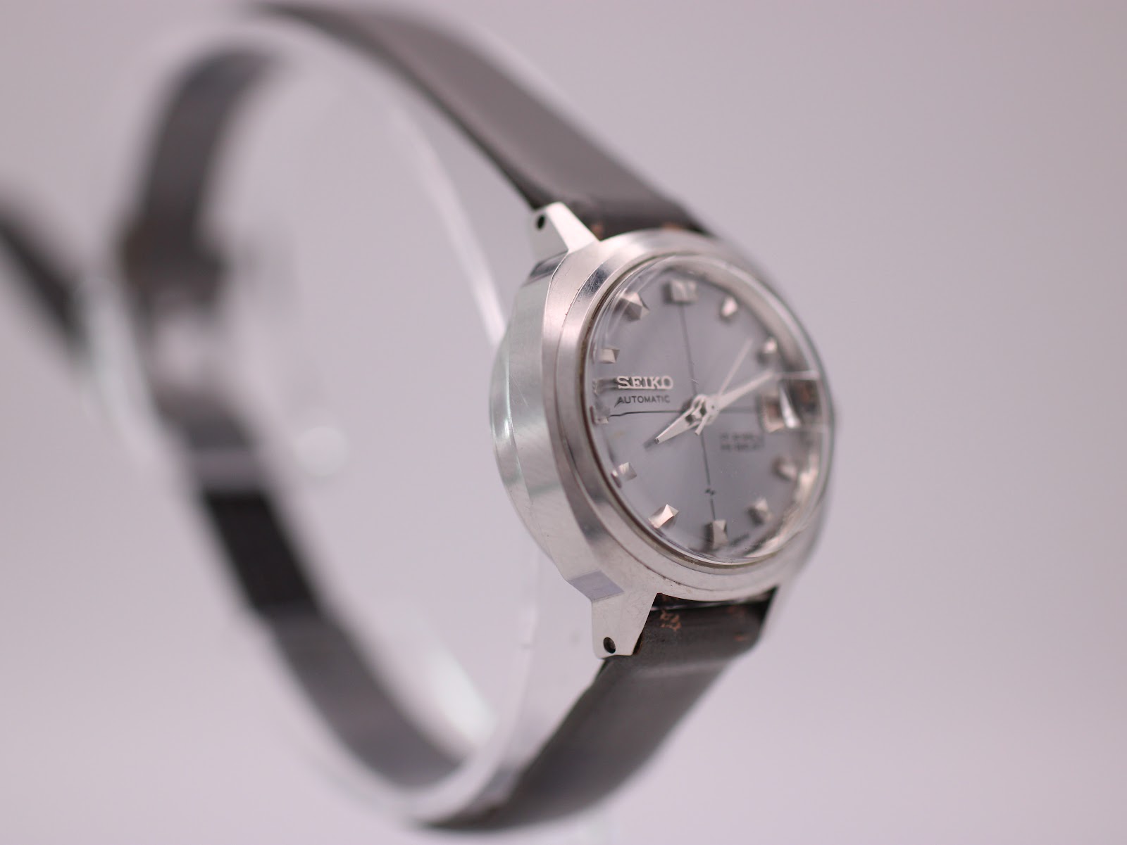 Angled side view of the Seiko watch on a stand, highlighting the polished stainless steel case and crown, with a grey leather strap visible.