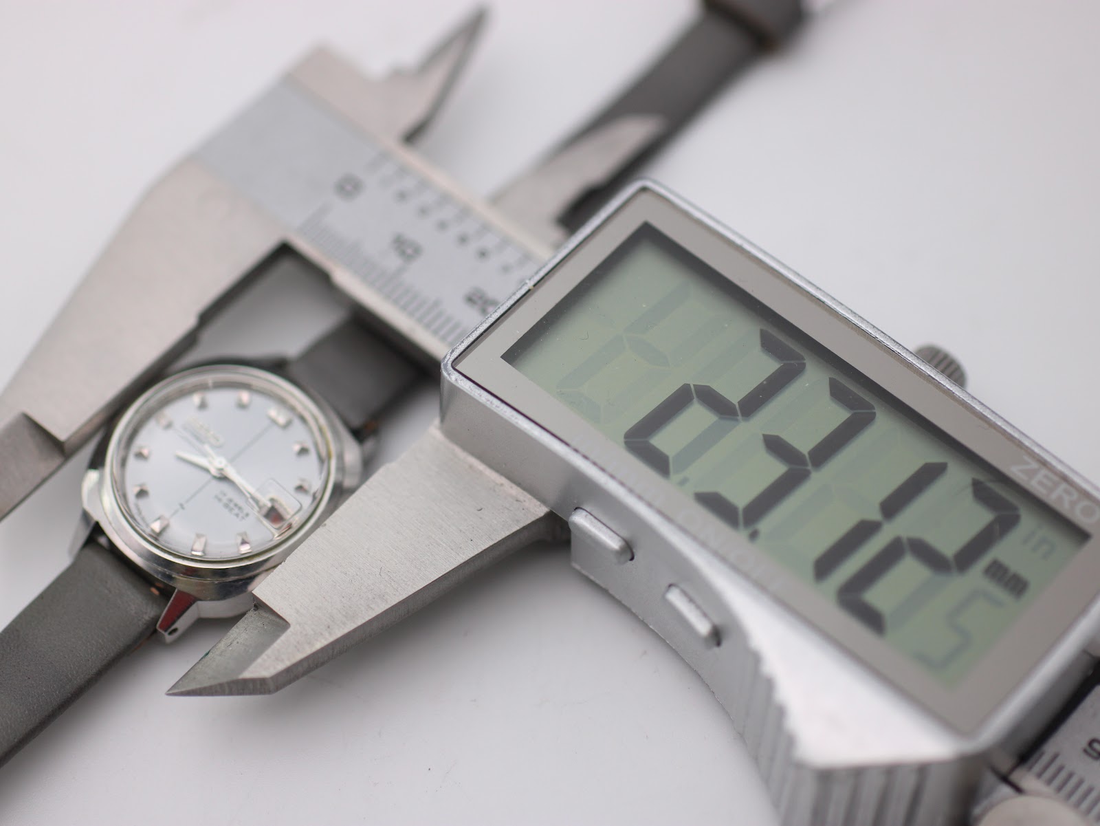 A digital caliper measuring the case diameter of the Seiko watch, with the display showing a precise reading of 27.12mm.