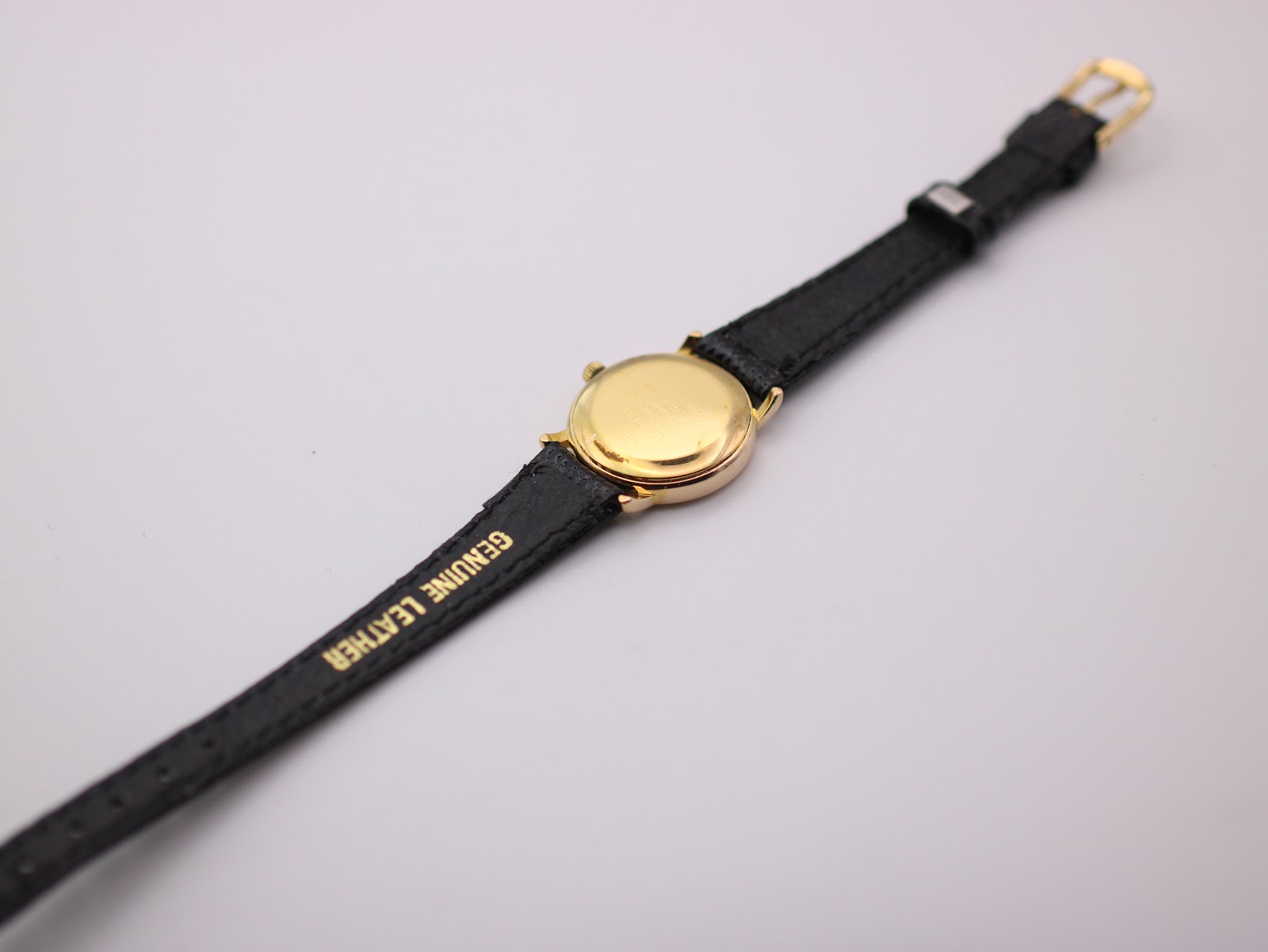 Back view of the Raymond Weil watch laid flat, showing the gold case back and "GENUINE LEATHER" text on the strap.