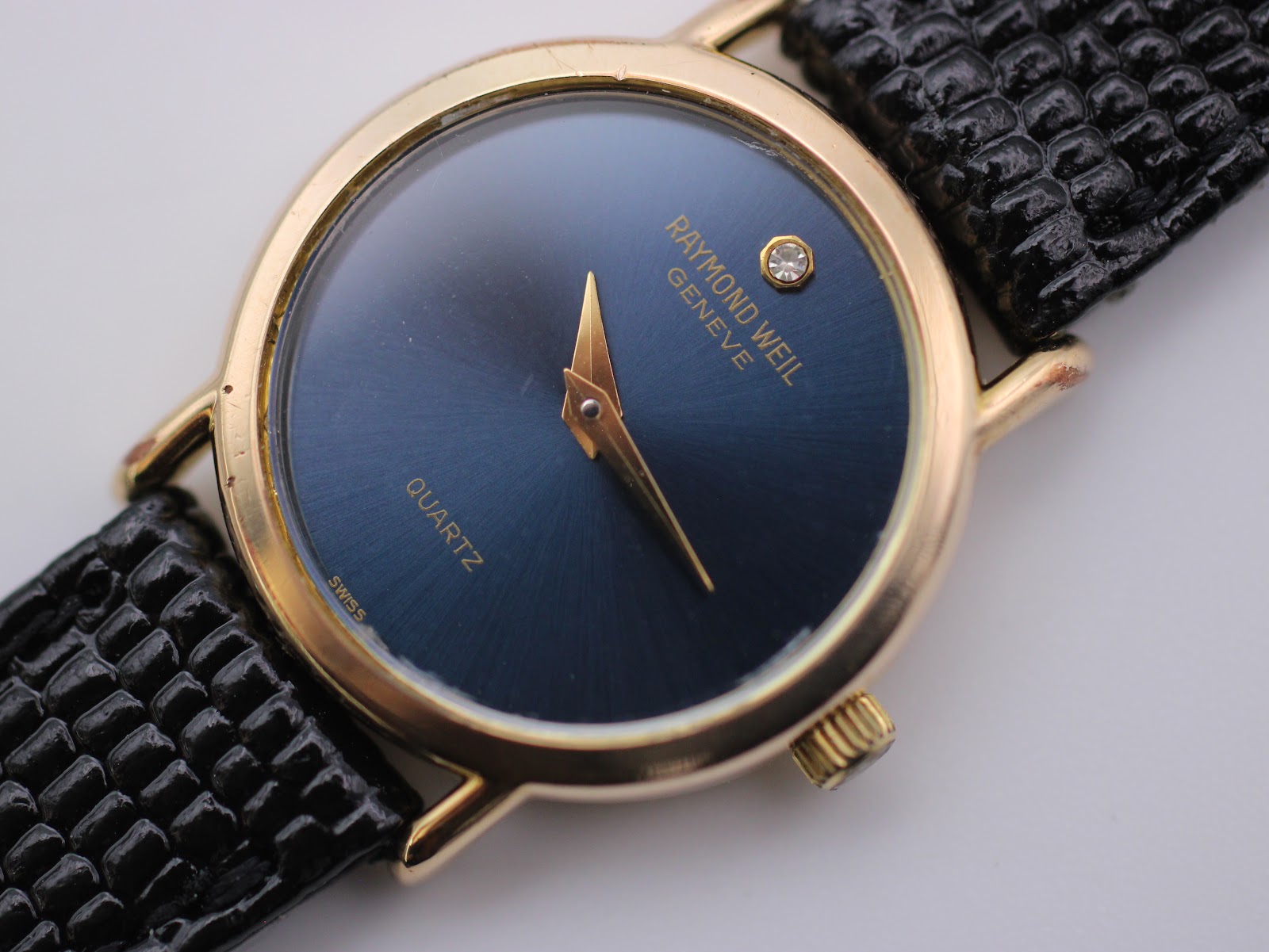 Close-up front view of the Raymond Weil blue dial, gold hands, diamond marker, and "RAYMOND WEIL GENEVE QUARTZ SWISS" text.