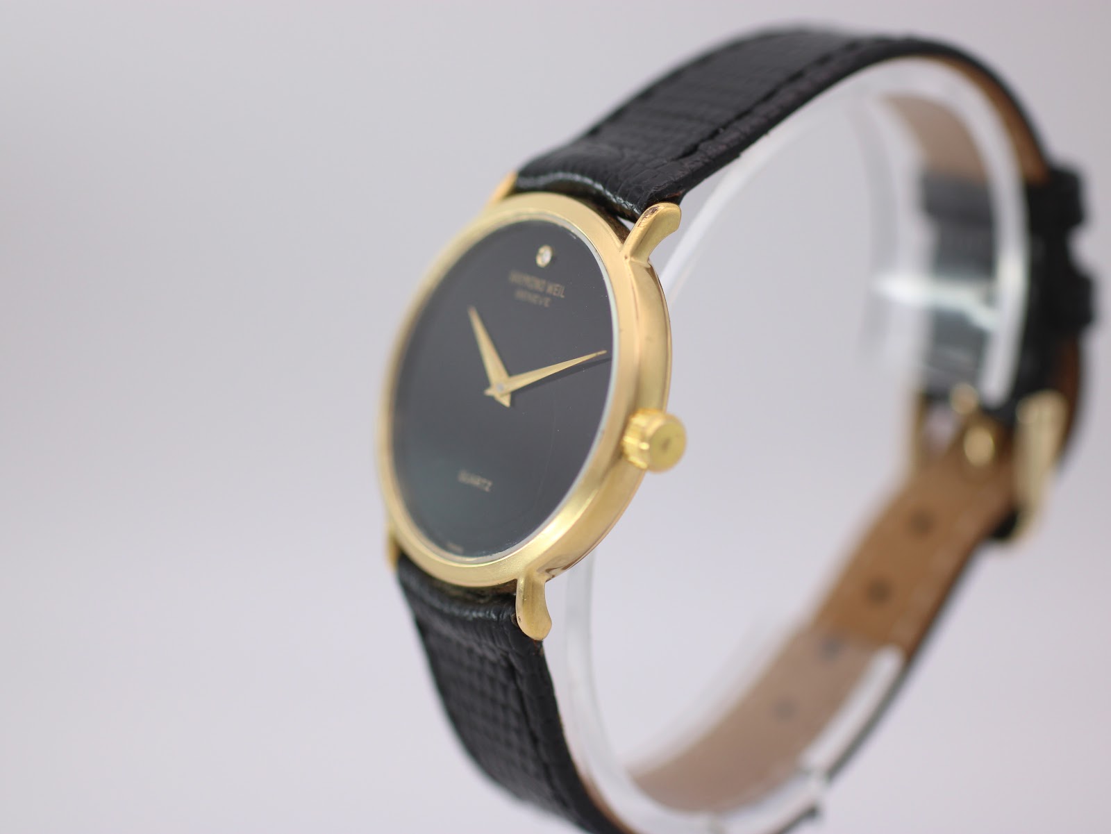 Angled side view of the Raymond Weil watch showing the gold case, crown, and black leather strap on a clear stand.