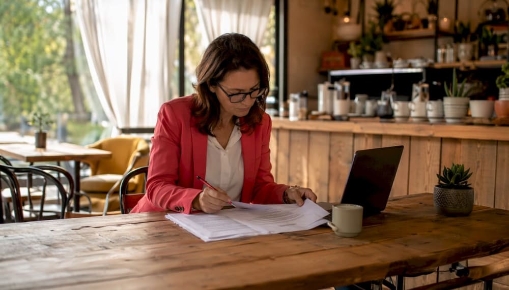 Femme annotant des documents administratifs dans un café, ordinateur portable à côté
