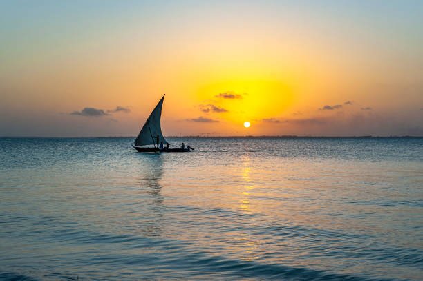 Traditional dhow sailing at sunset near Zanzibar coastline with orange sky reflections