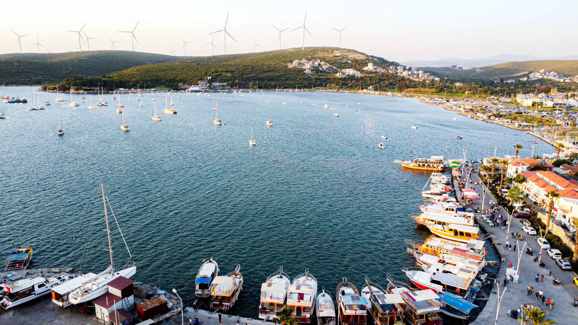 Le front de mer de Çeşme en fin d'après-midi, des bateaux de pêche et des bateaux de plaisance amarrés aux côtés de restaurants avec des tables en plein air