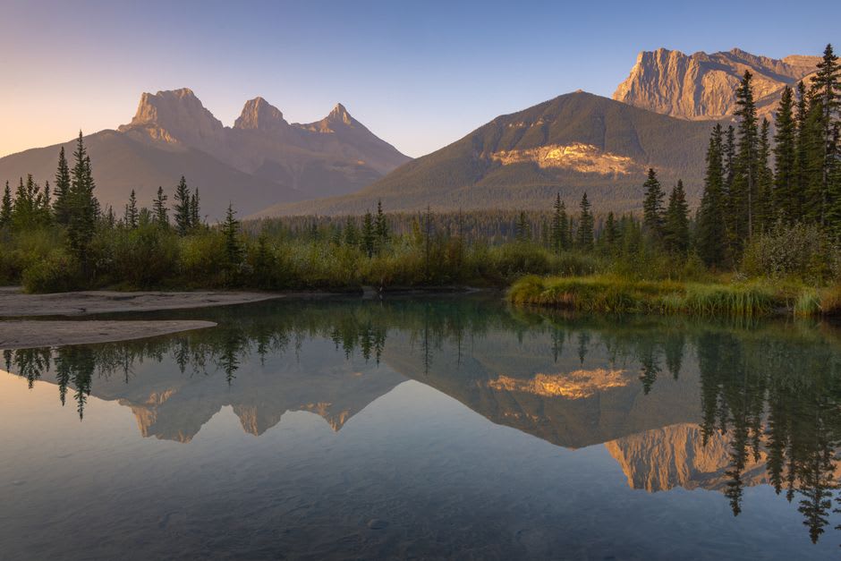 The Three Sisters peaks rising sharply above Canmore's rooftops at golden hour, autumn colour on the valley floor below