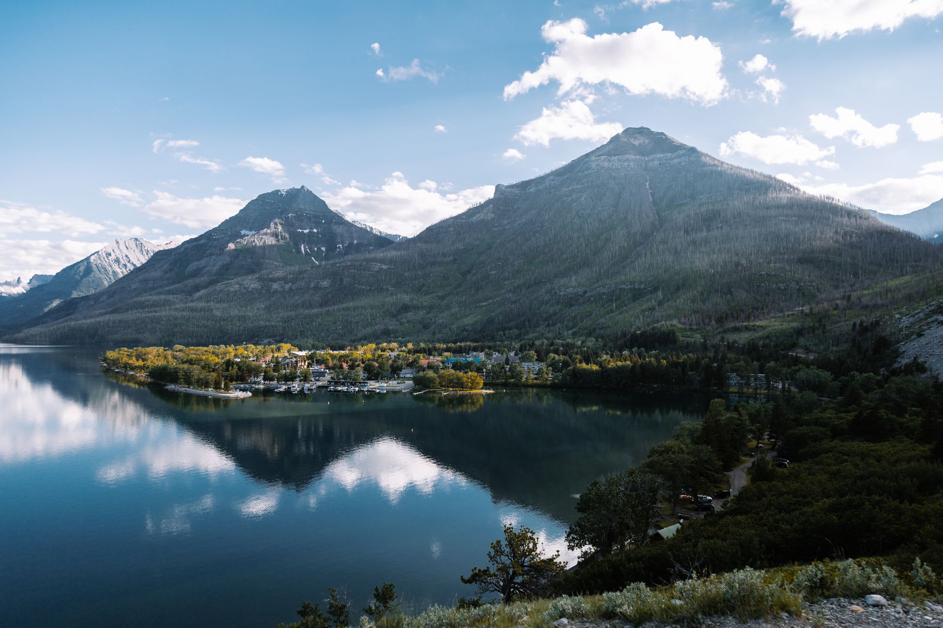 The Prince of Wales Hotel in late afternoon light, the wooden Victorian building on its green promontory reflected in the flat water of Upper Waterton Lake below